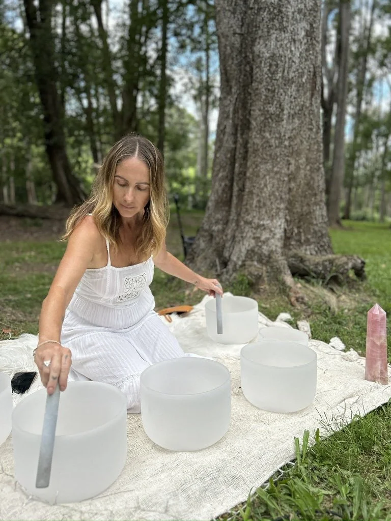 A woman in a white dress playing crystal bowls on a outdoor blanket in a forest setting.