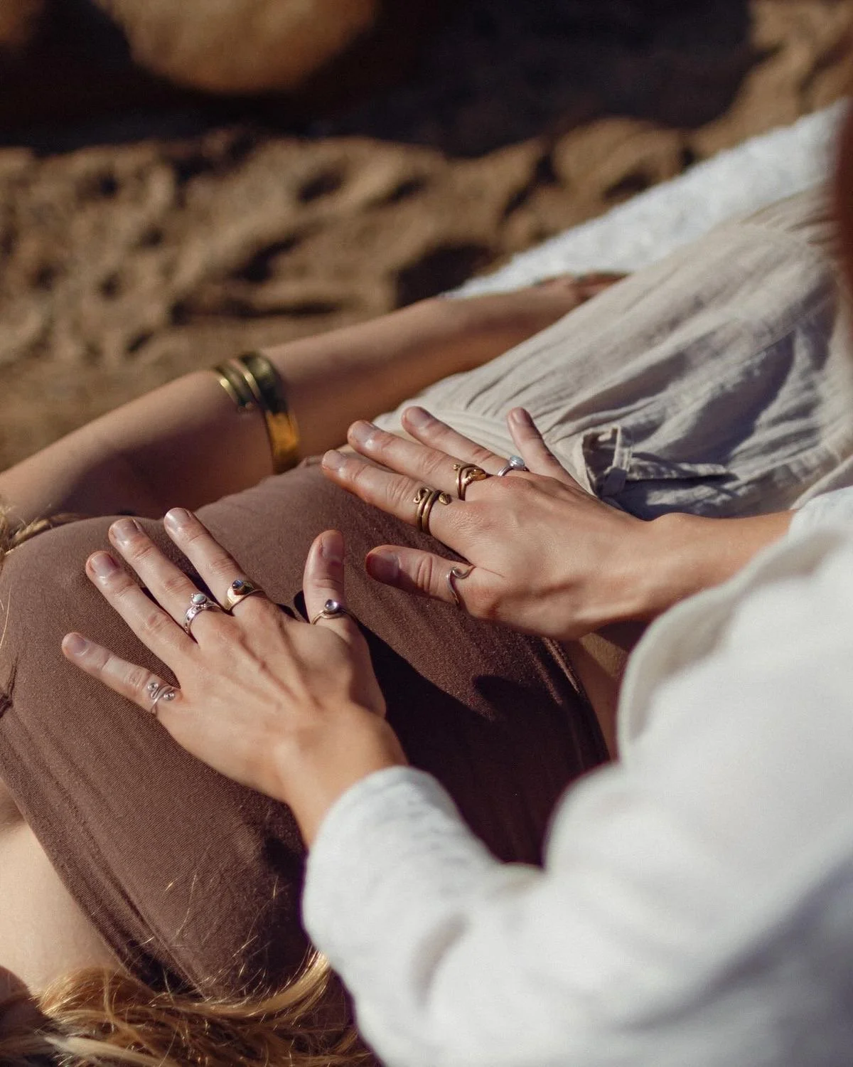 Two individuals are shown with their hands resting on each other's lap, both wearing various rings and bracelets. The scene appears intimate and relaxed.