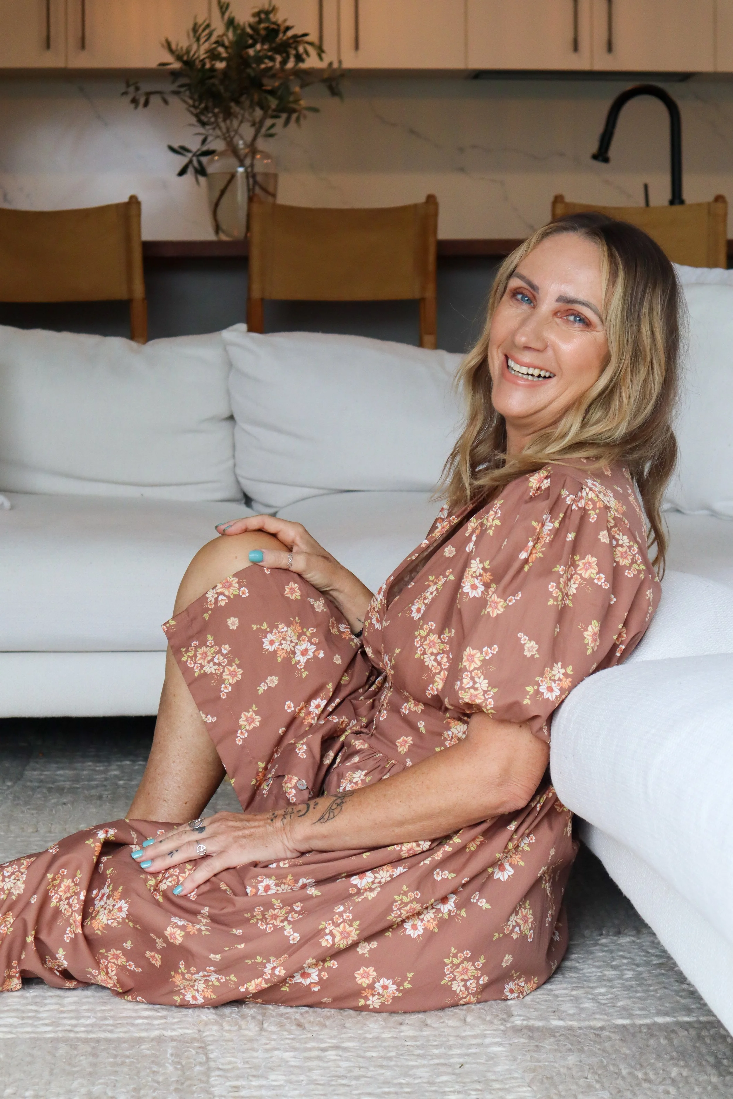 A woman sitting on the floor next to a white sofa, smiling and looking at the camera, wearing a floral dress, with a living room background.