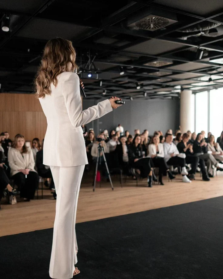 A woman in a white suit giving a presentation to a seated audience in a conference room.