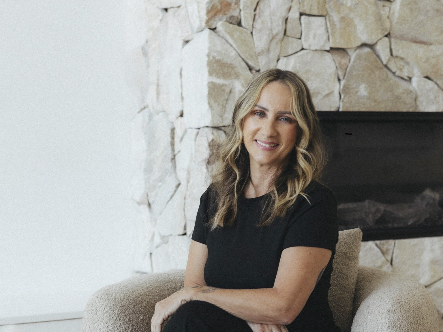 A smiling woman with wavy blond hair sitting on a beige armchair in front of a stone fireplace.