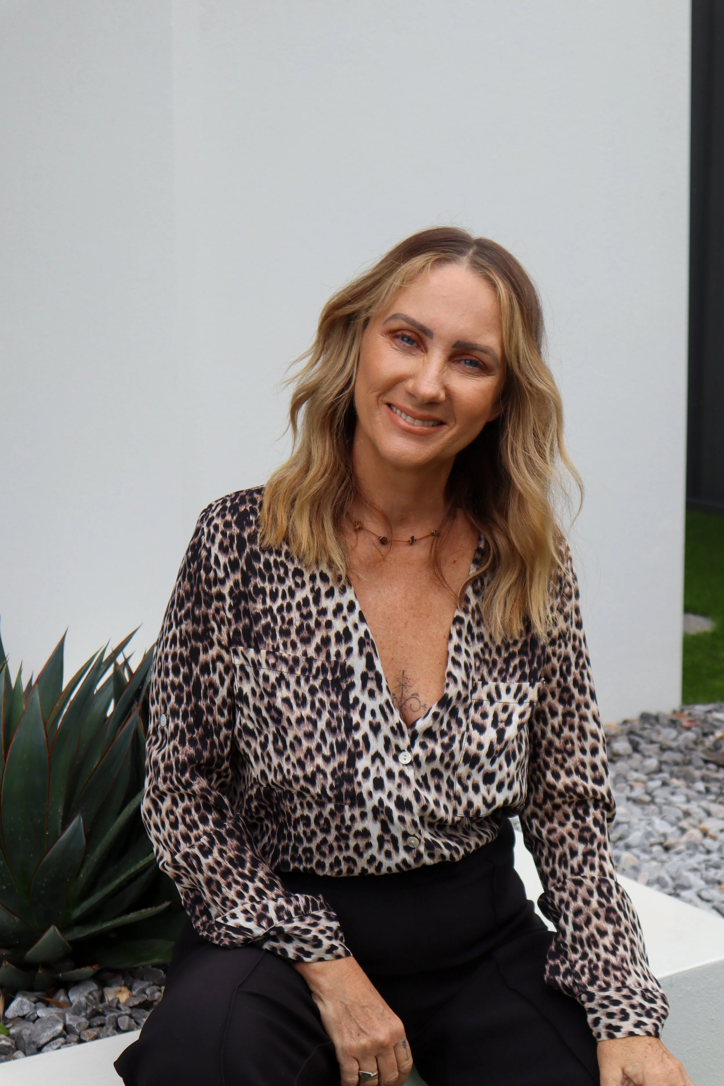 A woman sitting outdoors next to a large green plant with long pointed leaves, wearing a leopard print blouse and black pants, smiling at the camera.