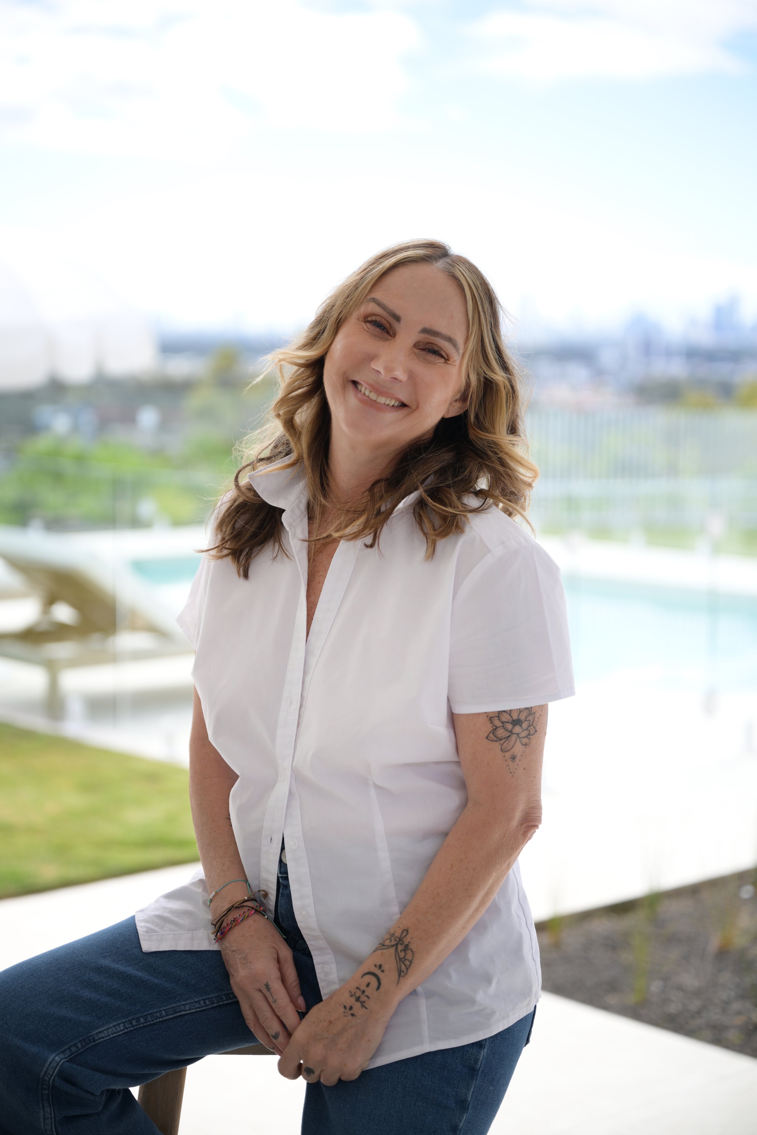 A smiling woman with shoulder-length blonde hair, wearing a white short-sleeve button-up shirt and blue jeans, sitting outdoors with a blurred background of a swimming pool and city skyline.