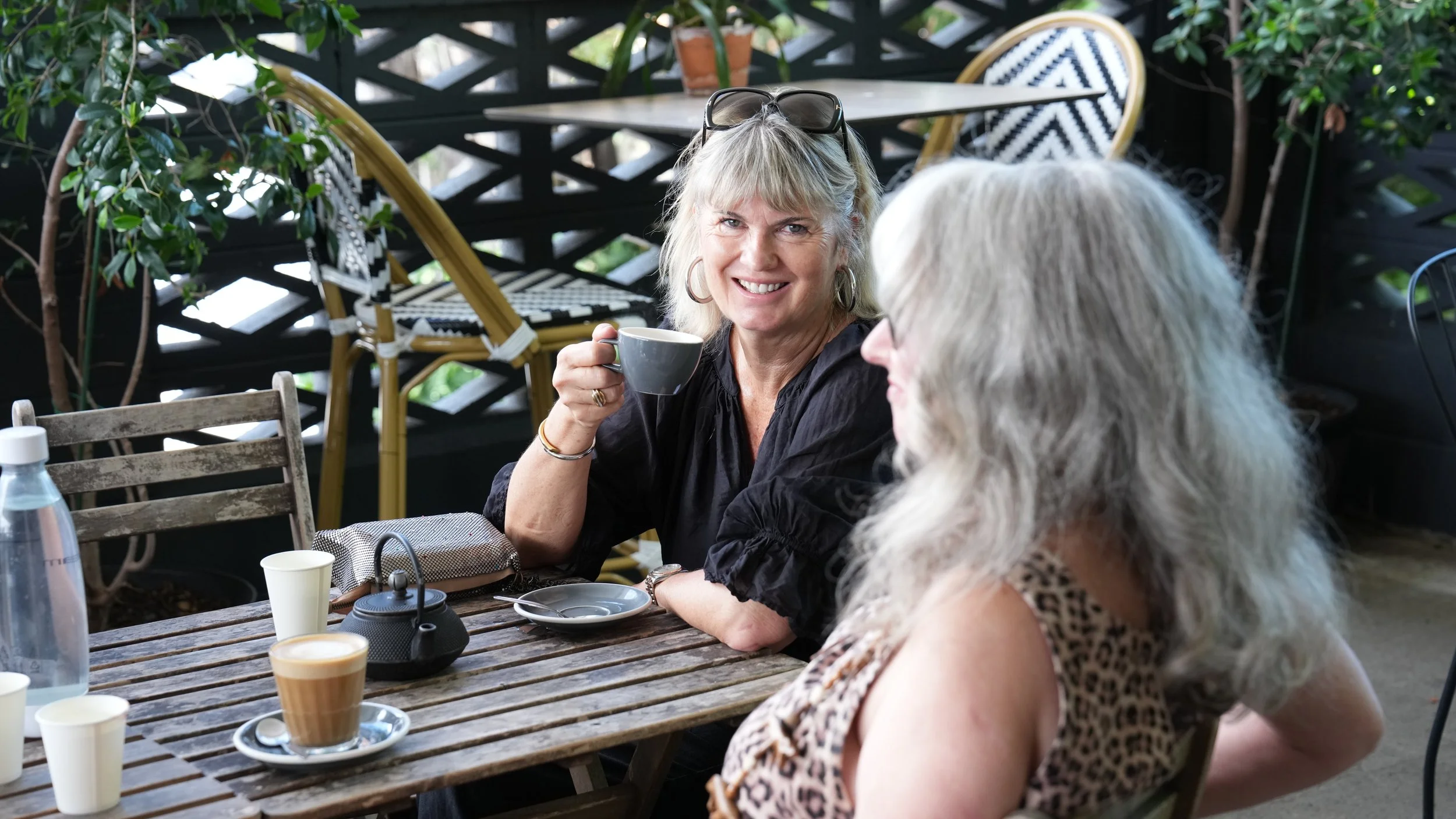 Two women with long, gray hair are sitting at a wooden outdoor table, having coffee or tea. The woman in the background is smiling and holding a gray mug, wearing black clothing and sunglasses on her head. The other woman is turned away, wearing a sleeveless leopard print top. On the table are coffee cups, a glass water bottle, a teapot, and an empty plate, with greenery and outdoor furniture in the background.