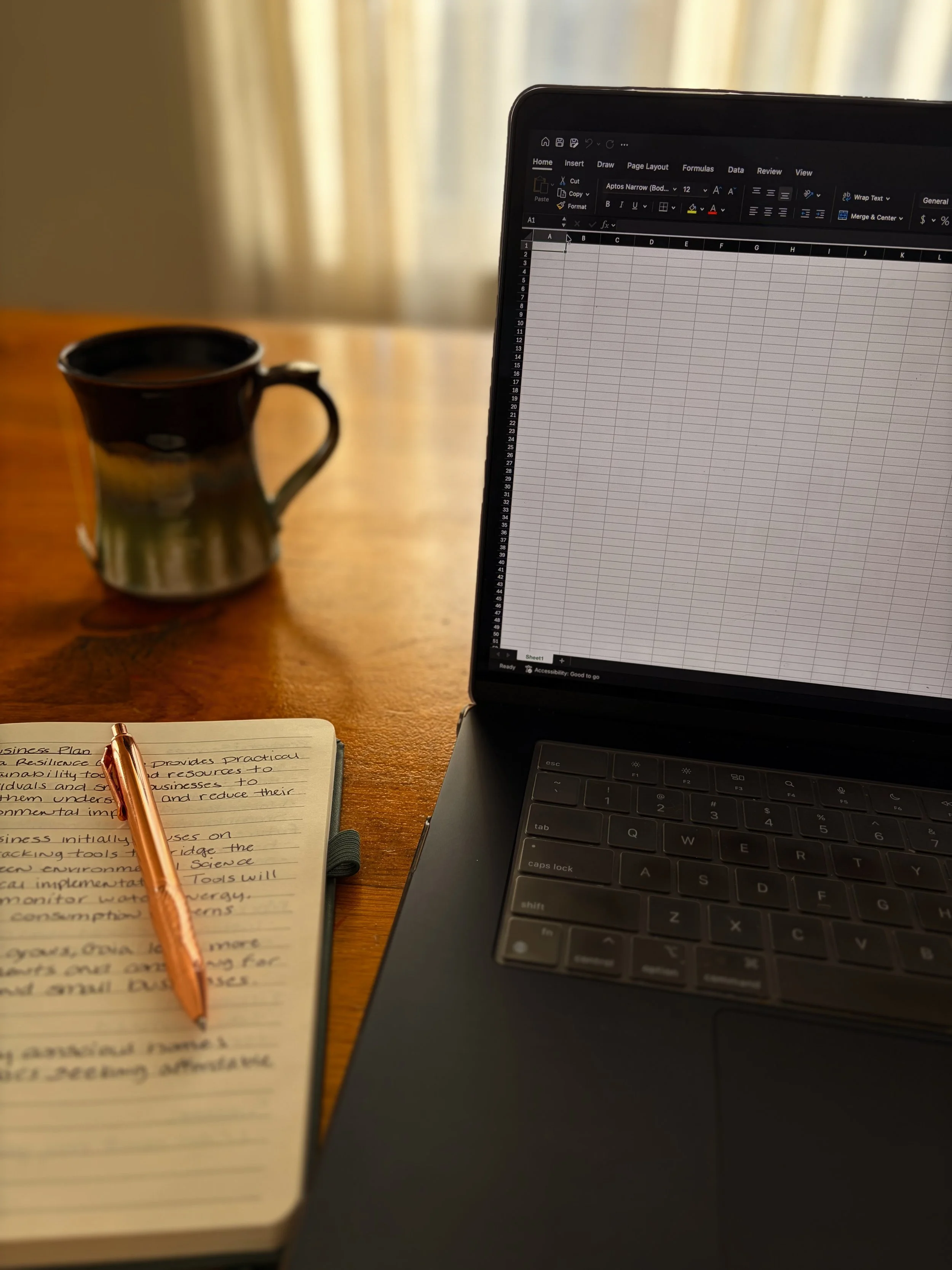A laptop with a spreadsheet open and a notebook with handwritten notes on the left hand side of the image. The notebook and laptop sit on a  wooden table, with a coffee mug in the background.