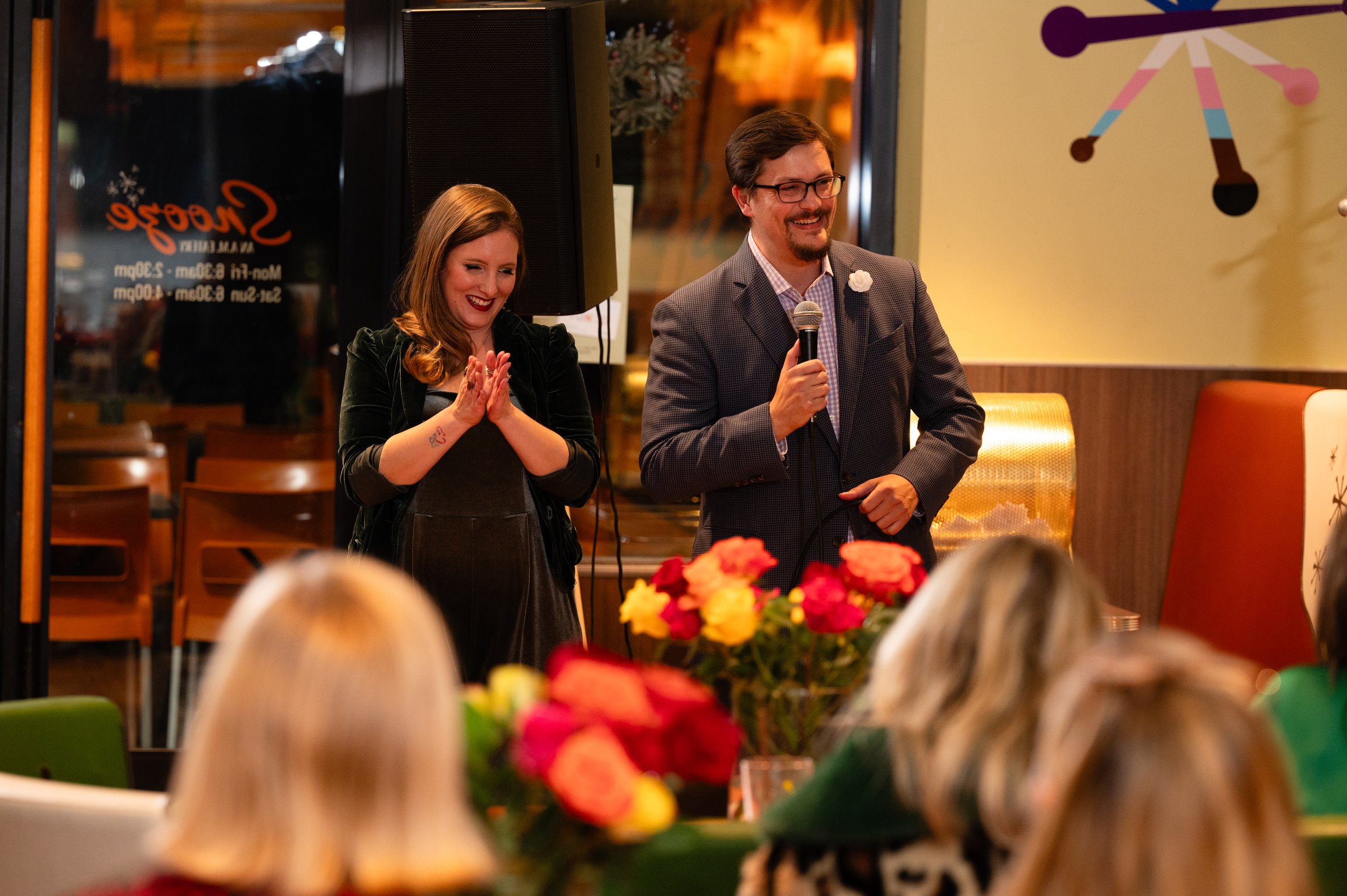 A man and woman speaking at an event in a restaurant or cafe. The woman has red hair, is wearing dark clothing, and is smiling with hands clasped. The man has glasses, facial hair, is wearing a checked shirt and blazer, and is holding a microphone, smiling. There are flowers on the table in front of them and audience members are in the foreground. The background shows a wall with abstract art and a window with a visitor sign.