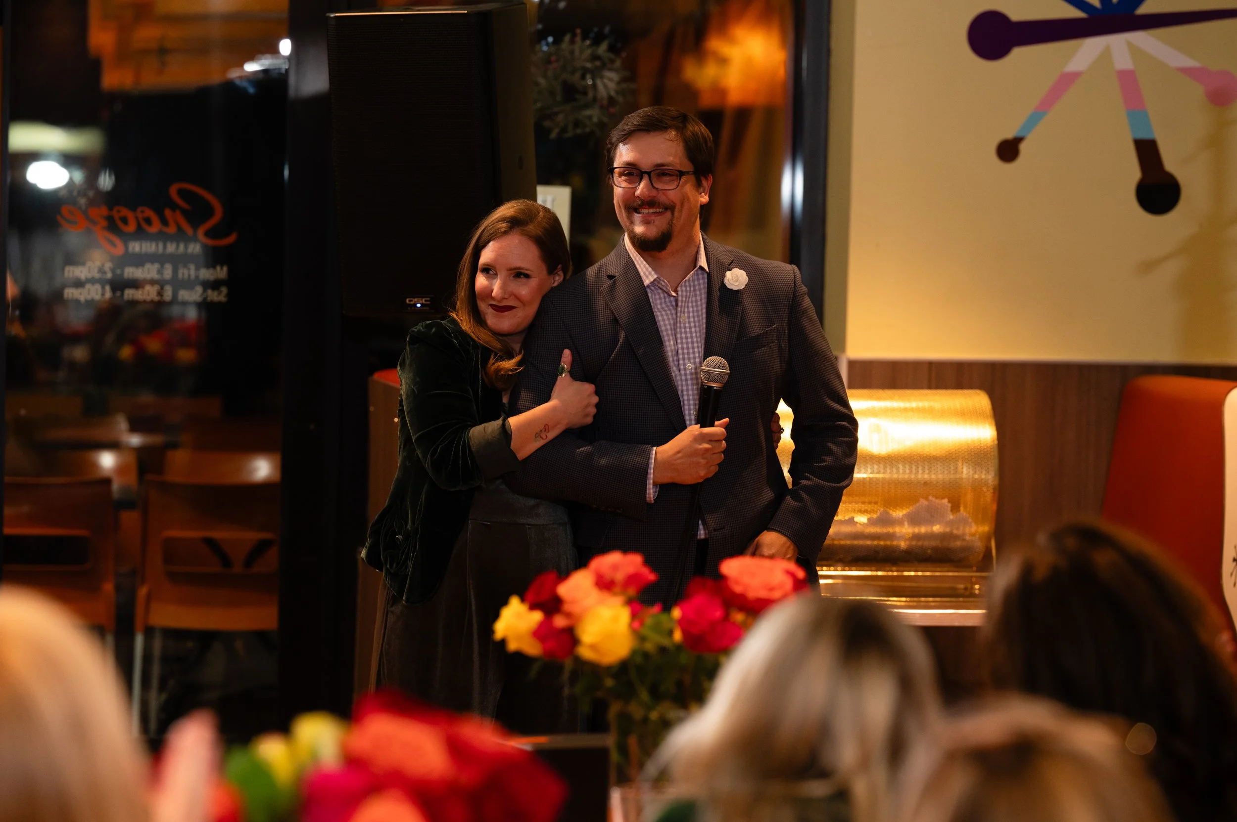 A woman leaning on a man holding a microphone, both smiling, at a social event inside a restaurant or bar, with flowers in the foreground and a decorated wall in the background.
