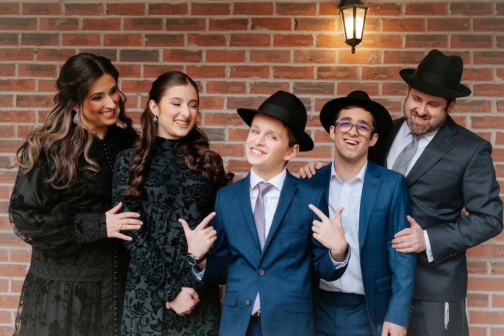 A group of five people, three women and two men, dressed in formal attire, standing against a brick wall, smiling and posing for a photo.