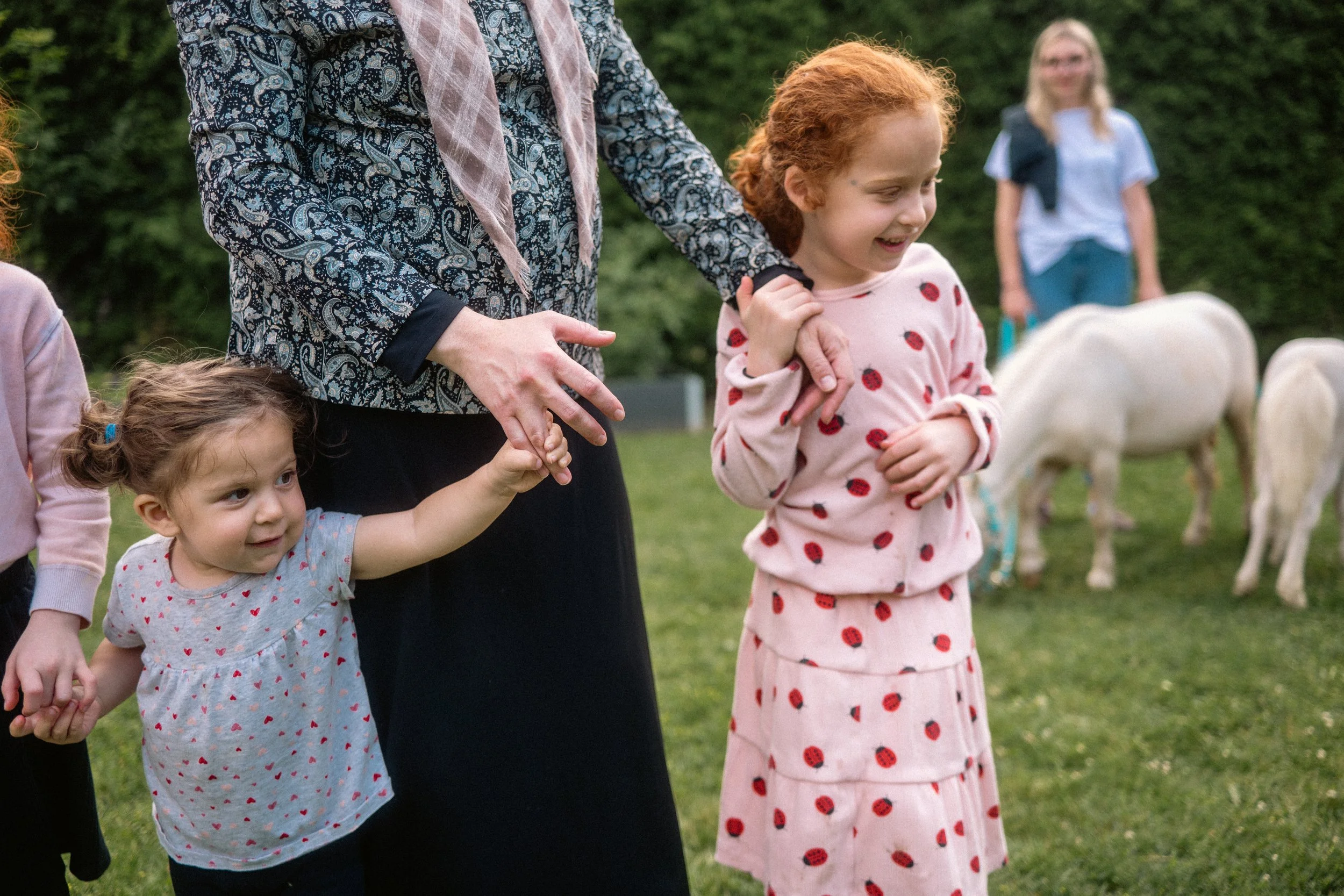 A woman and three young girls outside on the grass, with two white sheep nearby. The woman is holding the hand of a young girl with curly brown hair, wearing a grey dress with red hearts. Another girl with red hair, wearing a pink dress with red dots, is smiling and holding her hands together. In the background, a girl in a blue and white outfit stands near the sheep.