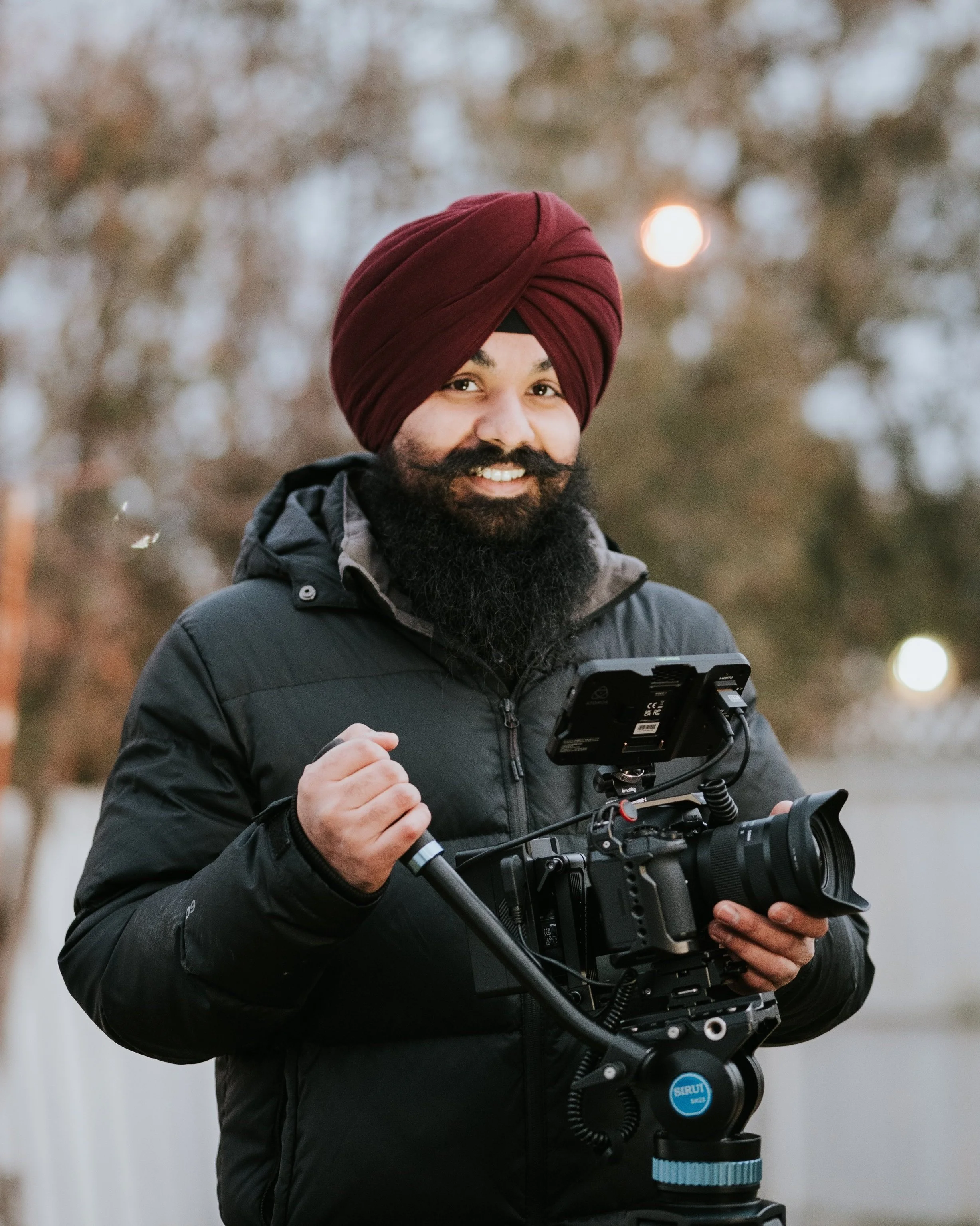 Man wearing a red turban and black jacket holding a camera with a stabilizer outdoors.