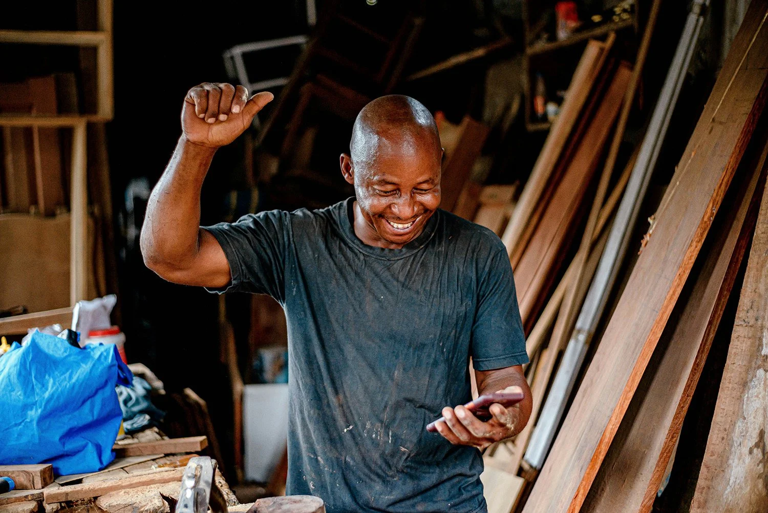 A man with a shaved head smiling and raising one fist in woodworking workshop, holding a phone in his other hand, surrounded by wooden planks and tools.