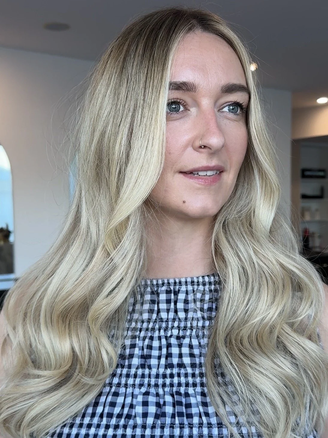 A woman with long, wavy blonde hair and blue eyes, wearing a black and white checkered top, indoors with a blurred background.