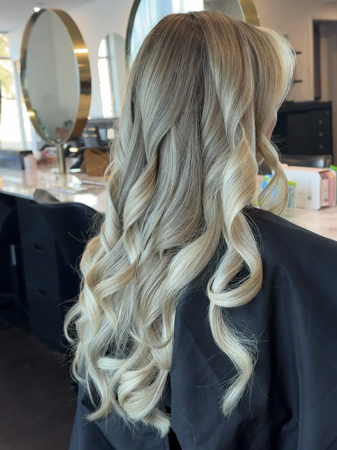 Woman with long, curled blonde hair in a salon with mirrors and hair products in the background.