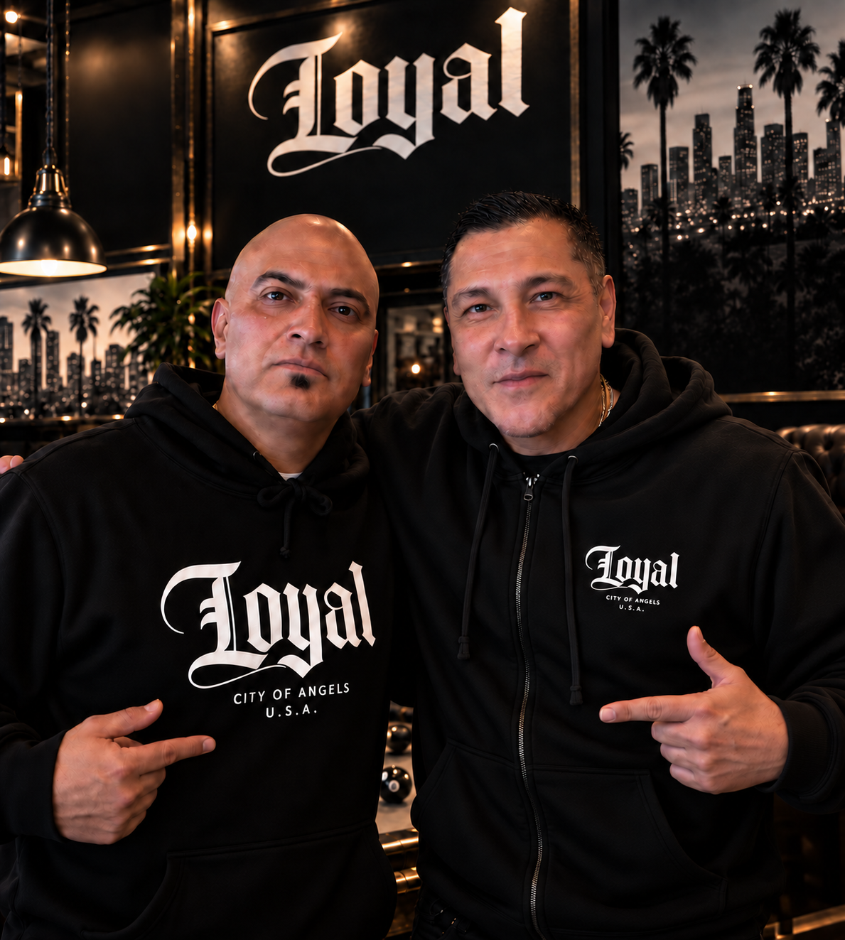 Two men wearing black hoodies with 'Loyal, City of Angels, U.S.A.' printed on them, standing in a dimly lit room with cityscape and palm tree artwork on the wall behind them. They are posing for the camera, with one pointing to his shirt.