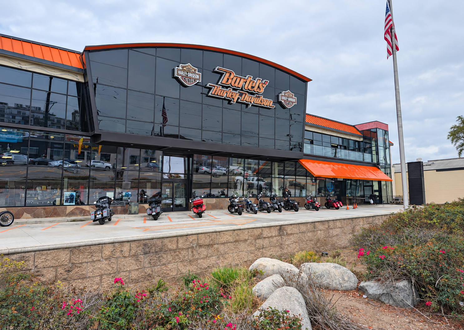Exterior view of a Harley-Davidson dealership building with a black and orange sign, multiple motorcycles parked in front, and an American flag. The building has large glass windows with cars visible inside and outside.