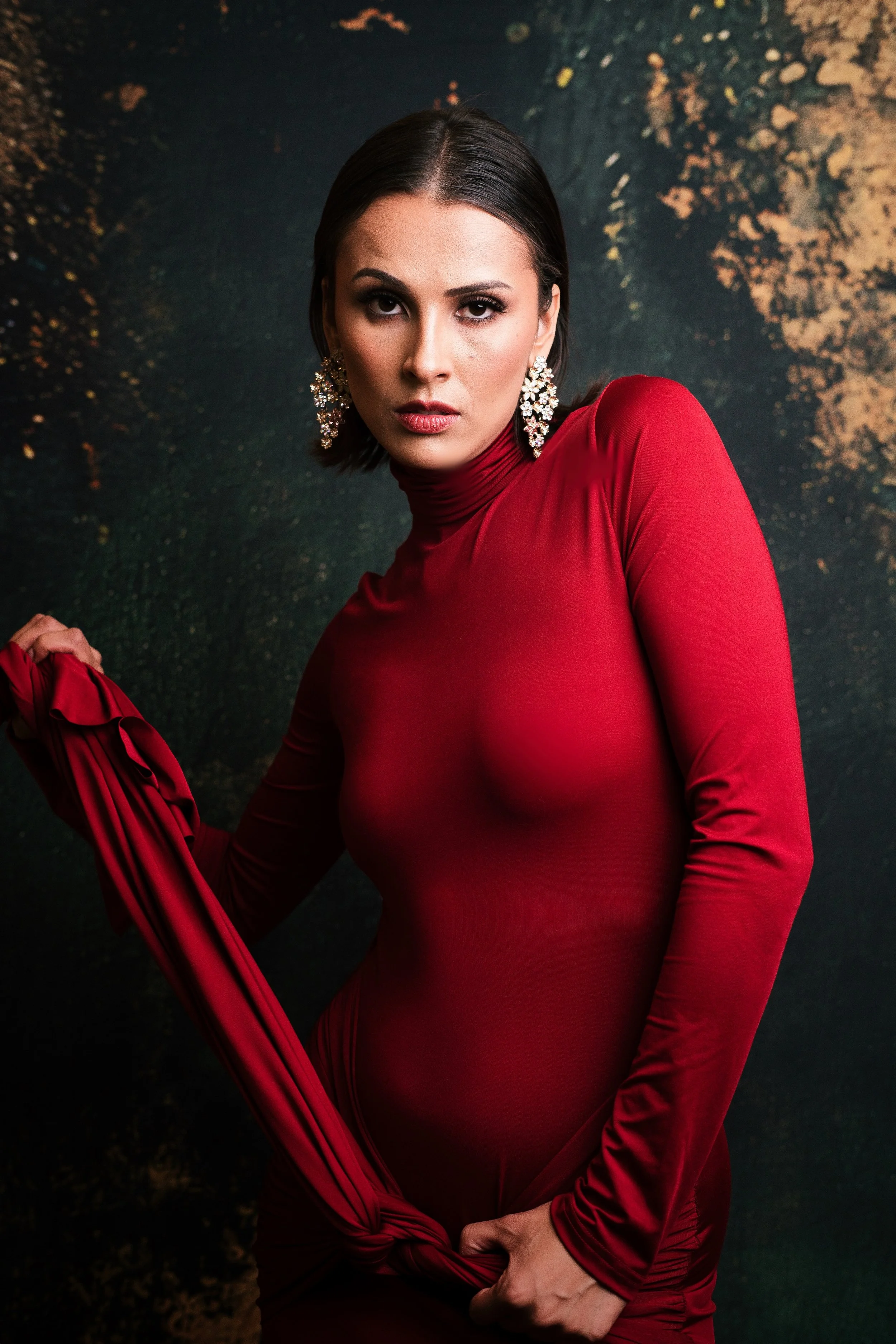 A woman with dark hair wearing a red long-sleeve turtleneck dress and ornate earrings, posing against a dark, textured background with gold accents.