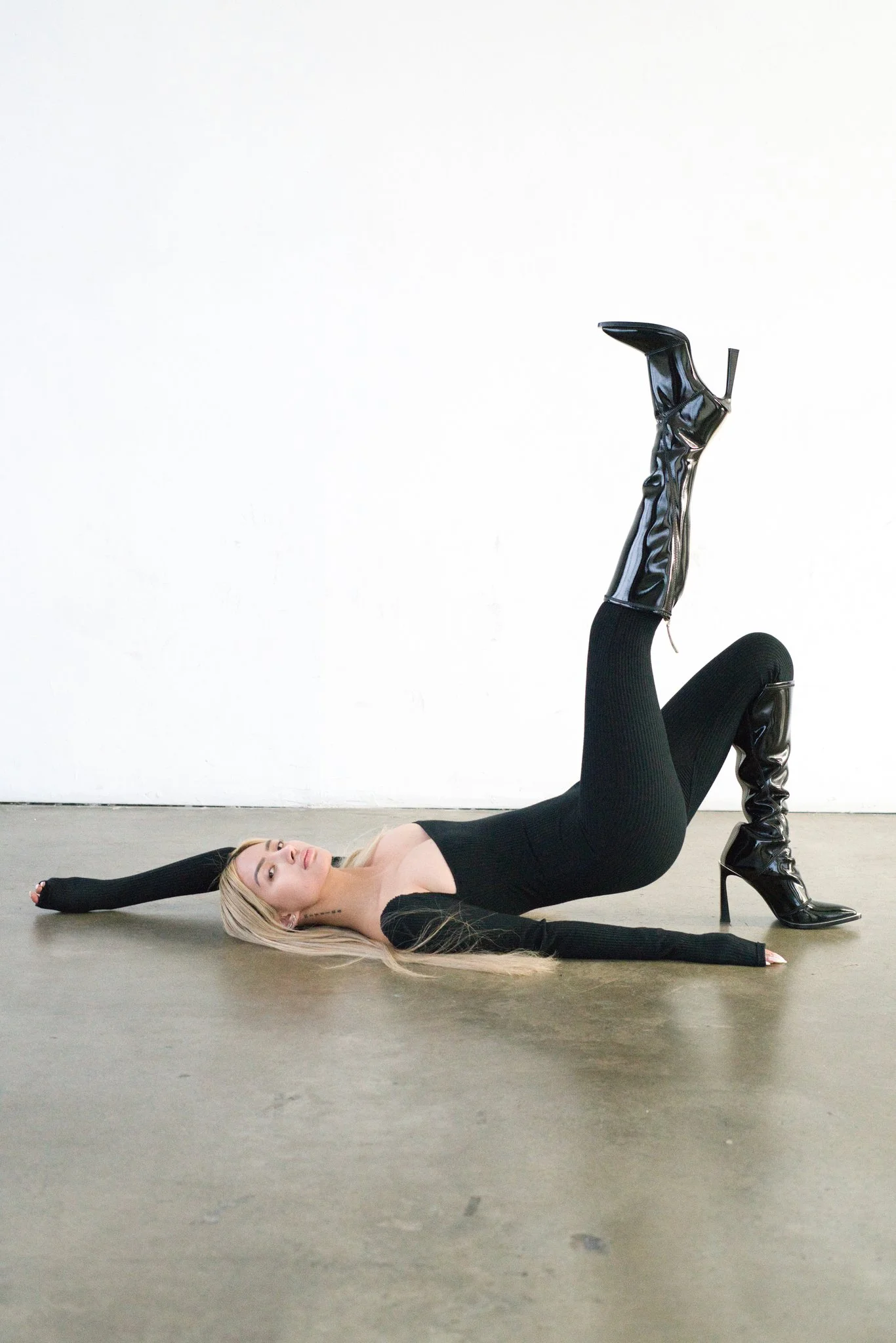 A woman lying on her back on a grey floor with one leg raised and bent, wearing black thigh-high boots and a black dress with long sleeves, in front of a white wall.