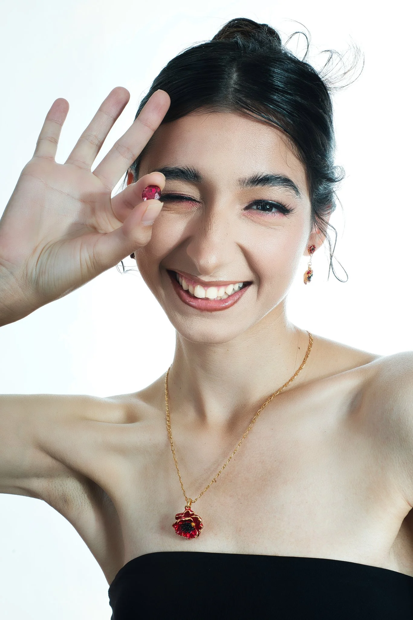 A woman with dark hair and a black strapless top smiling and making an OK gesture with her left hand, holding a pink ring near her eye, wearing a gold necklace with a red pendant and matching earrings.