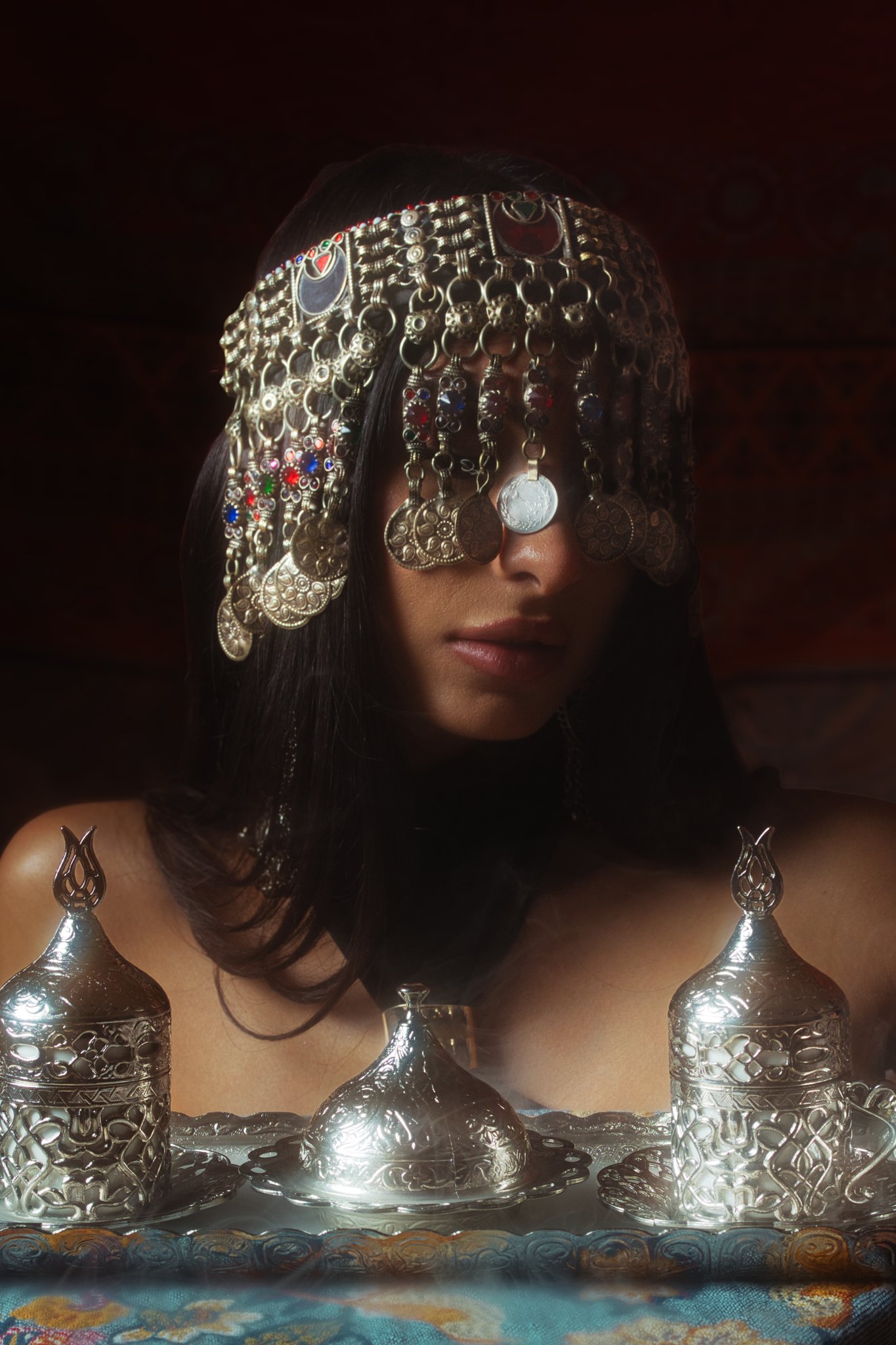 Woman with intricate silver jewelry and fabric headpiece, surrounded by silver objects on a patterned cloth.