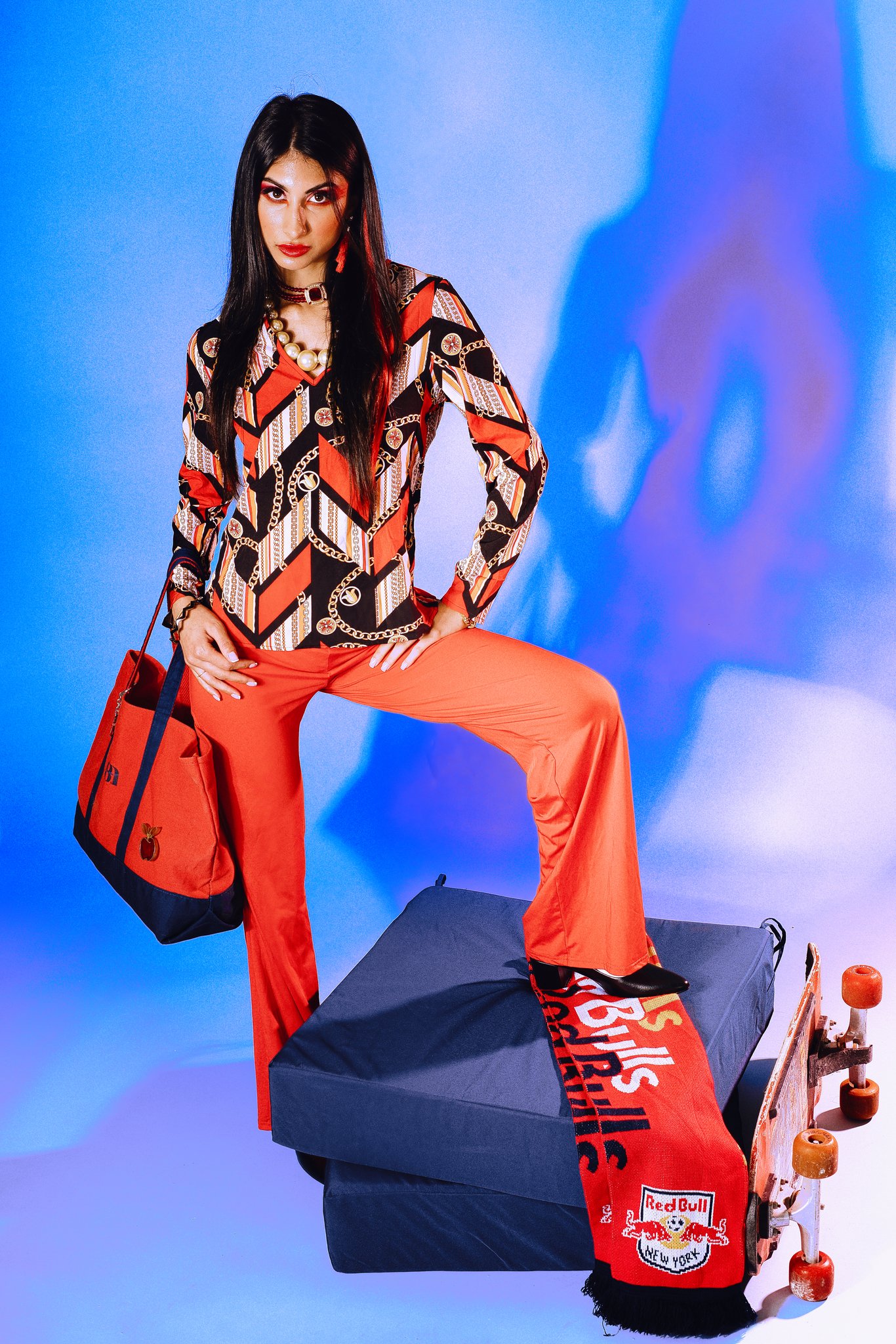 Young woman with long dark hair wearing a colorful patterned blouse, orange pants, and a pearl necklace. She stands with one foot on a skateboard and poses against a blue background, with Red Bull sports gear nearby.