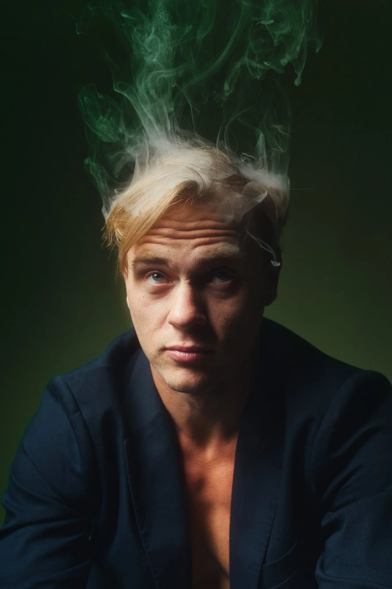 A young man with blond hair and blue eyes wearing a dark blazer, looking upward with smoke rising from his head against a green background.