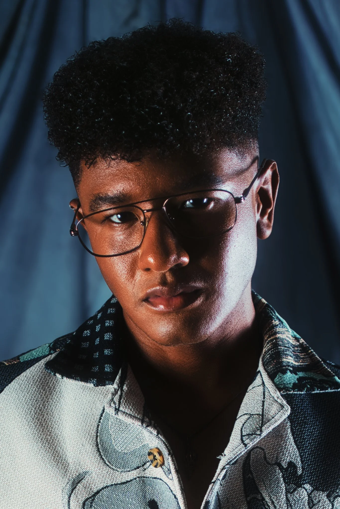 Close-up of a person with dark curly hair, wearing glasses, and a patterned shirt, against a backdrop of dark blue curtains.