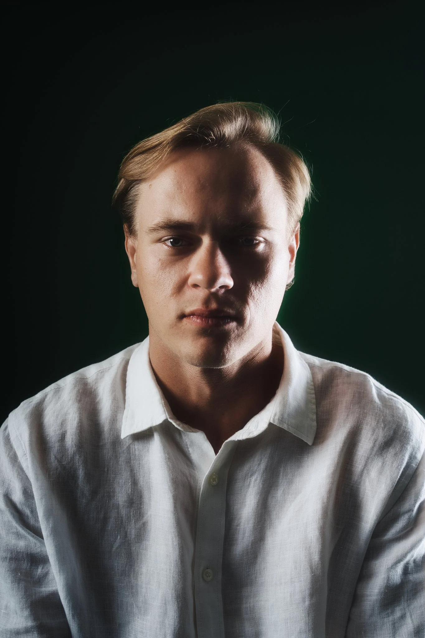 A young man with light brown hair, wearing a white shirt, looking directly at the camera with a serious expression, against a dark green background.