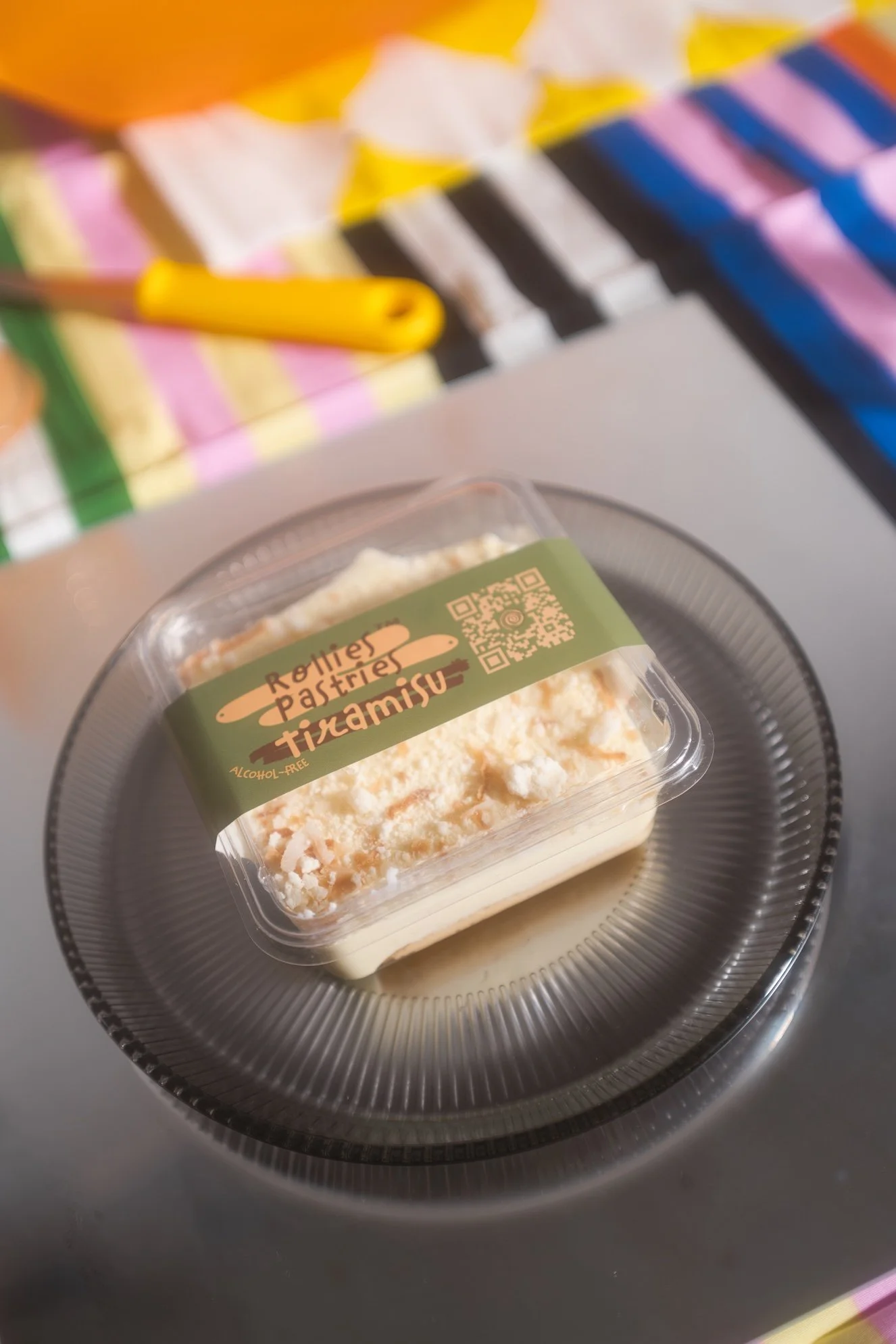 A packaged slice of vegan Tiramisu dessert on a silver tray, with a colorful, striped fabric in the background.