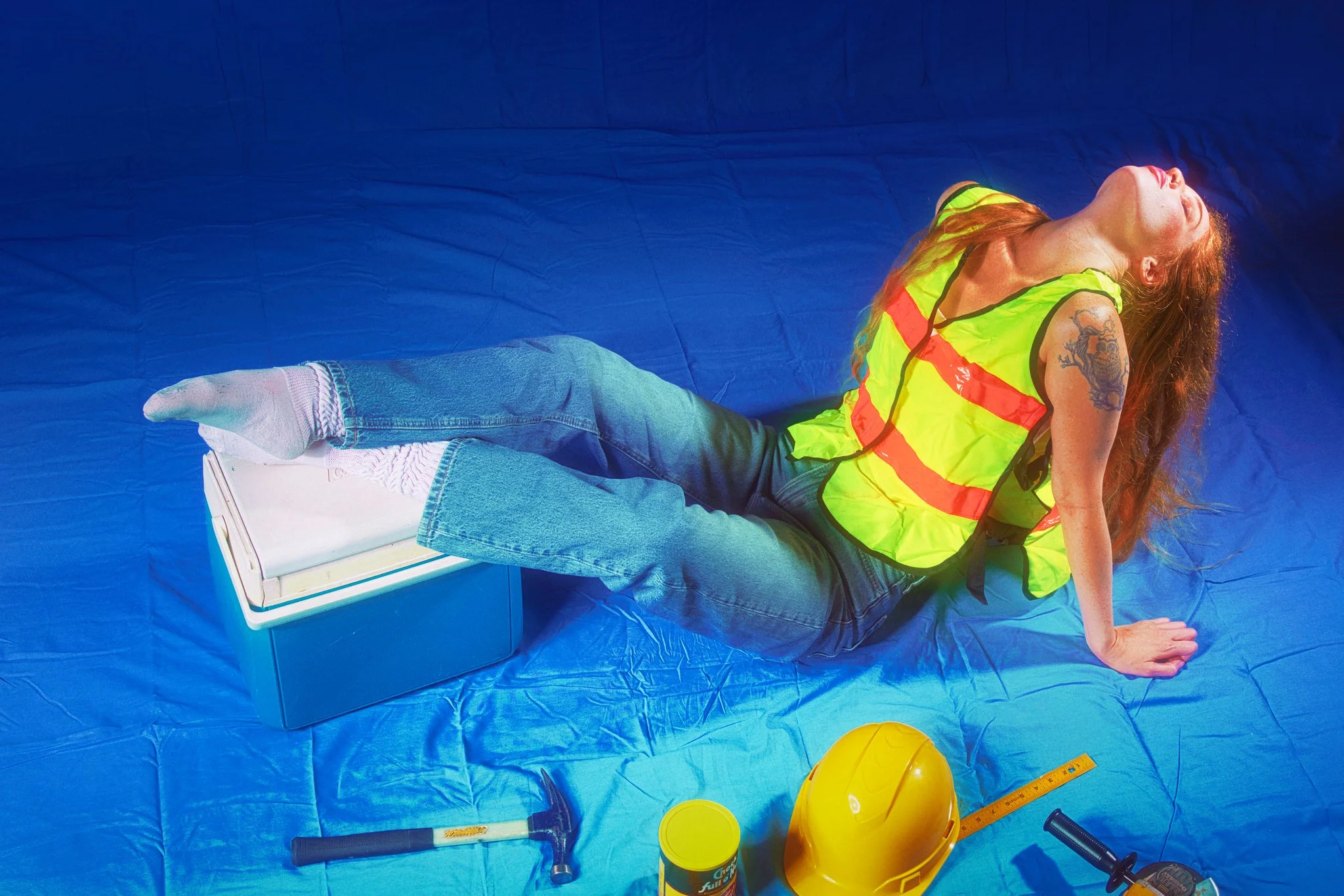 A woman in a yellow safety vest and jeans is lying on a blue tarp, with her head tilted back and eyes closed. Construction tools and a yellow hard hat are nearby.