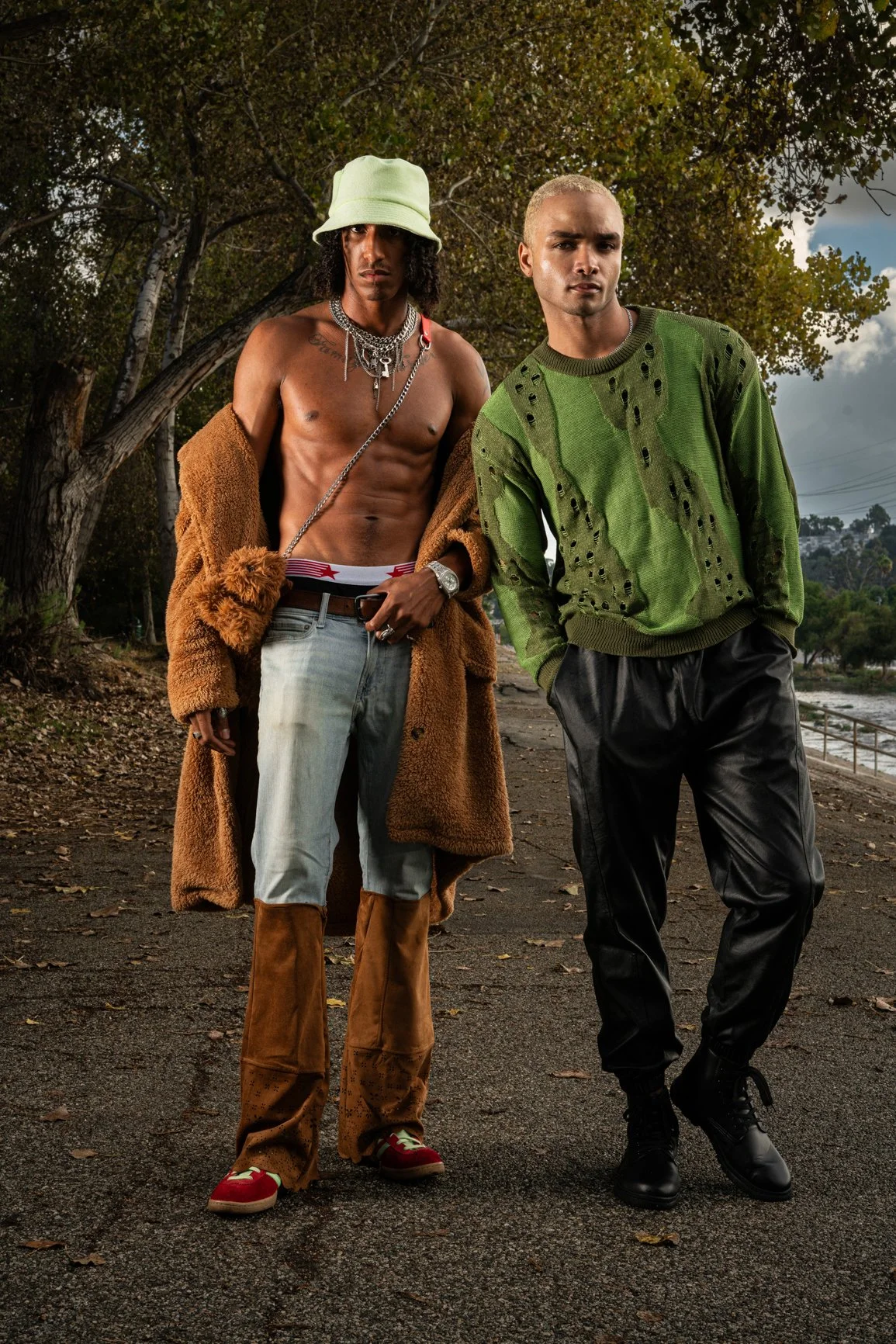 Two young men standing outdoors on a cloudy day. One is shirtless, wearing a green bucket hat, multiple necklaces, a brown coat hanging off his shoulders, distressed jeans, and red sneakers. The other is wearing a green ripped sweater, black pants, a