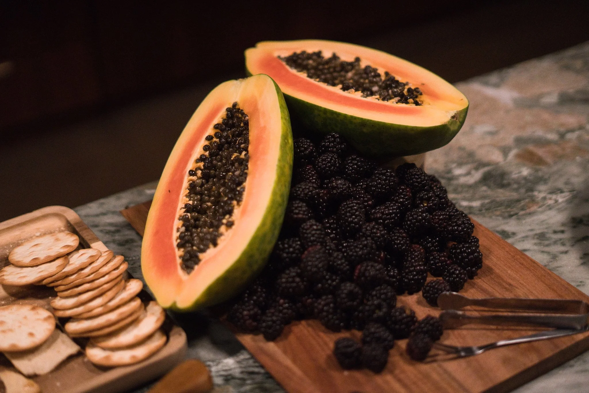 Halved papaya with black seeds on a wooden platter, surrounded by blackberries and crackers, on a marble surface.