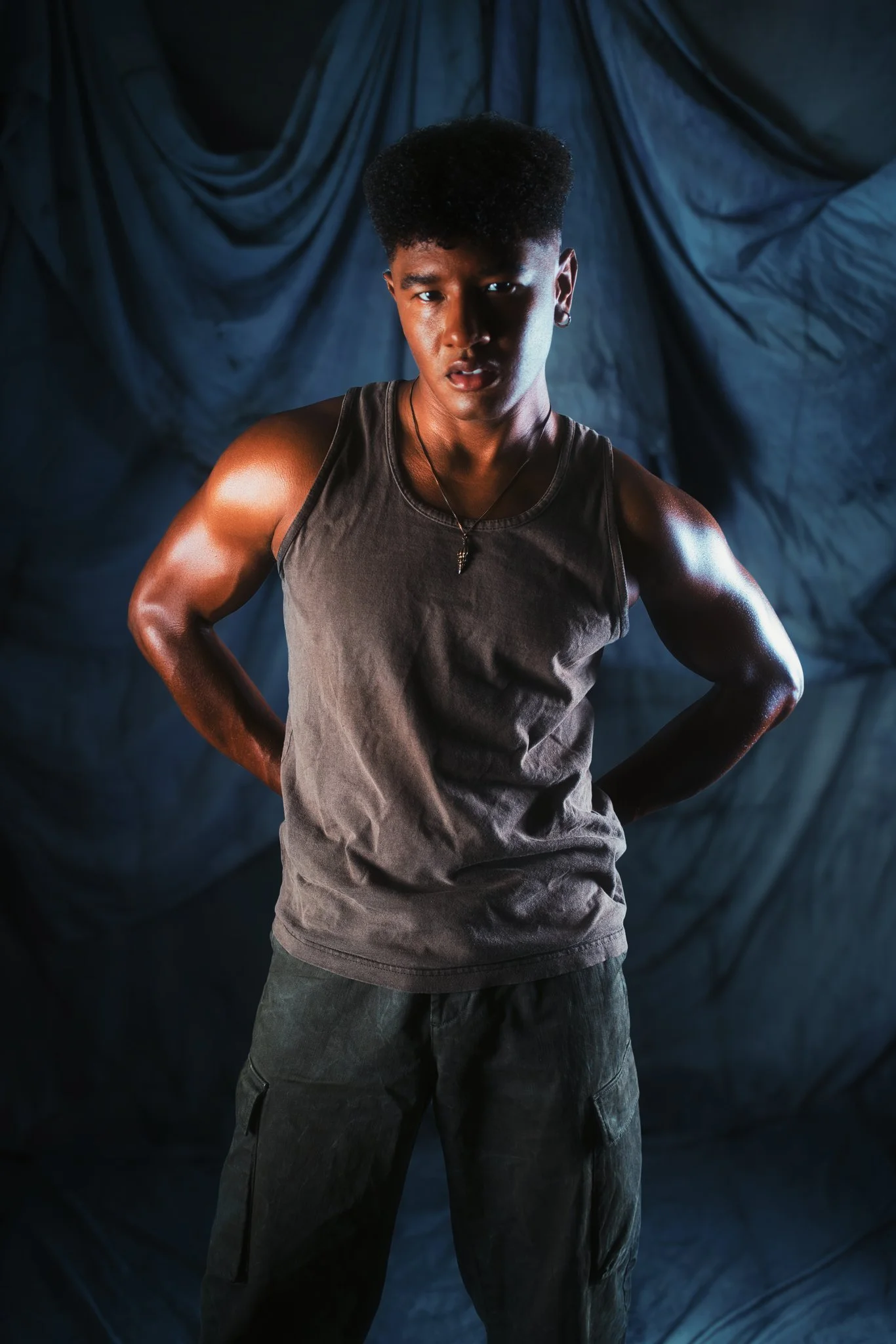 A person with short curly hair, wearing a sleeveless shirt and cargo pants, standing with hands on hips against a dark, draped background.