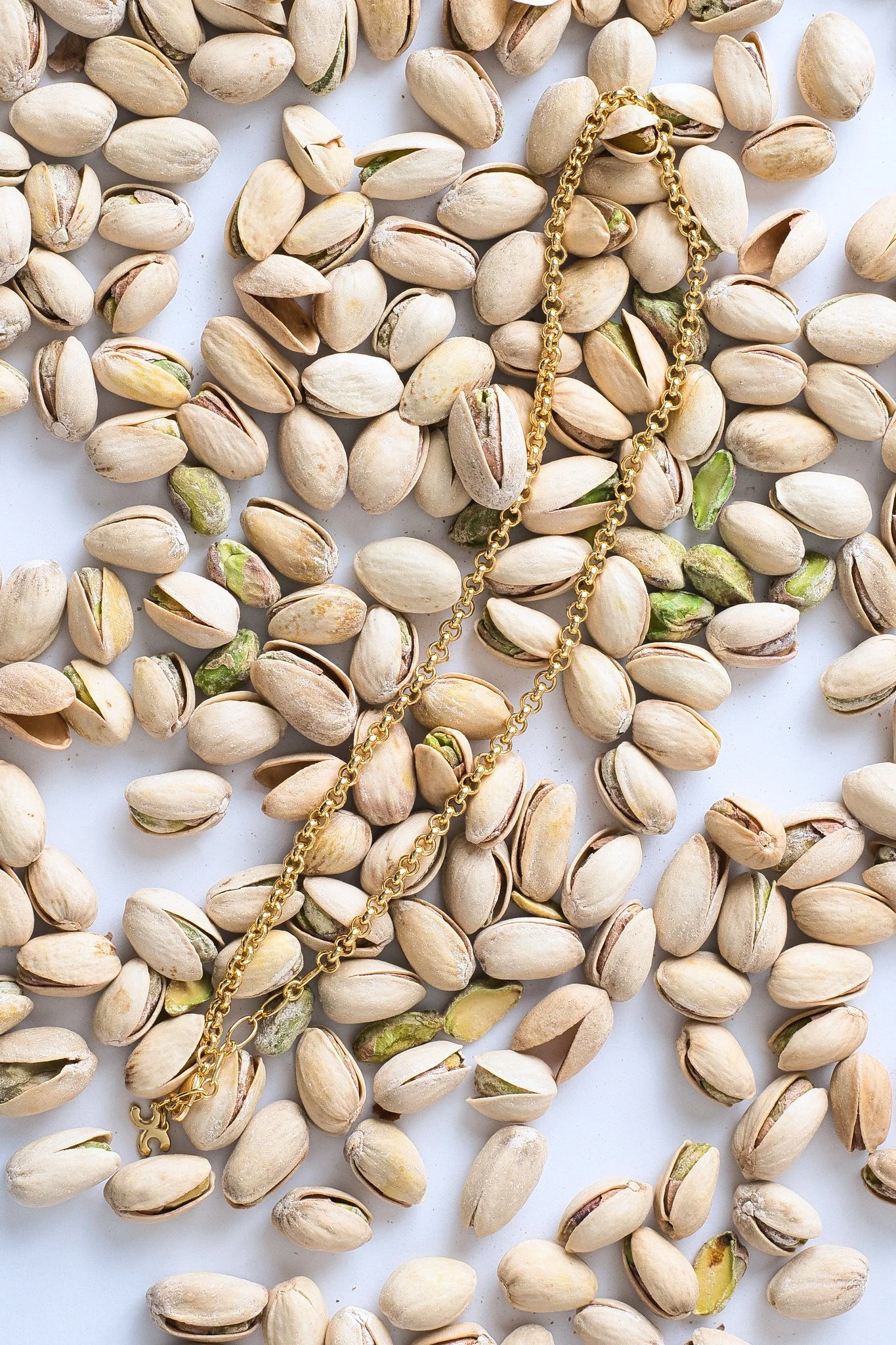 Close-up of pistachio nuts scattered on a white surface, with a gold chain necklace resting among them.
