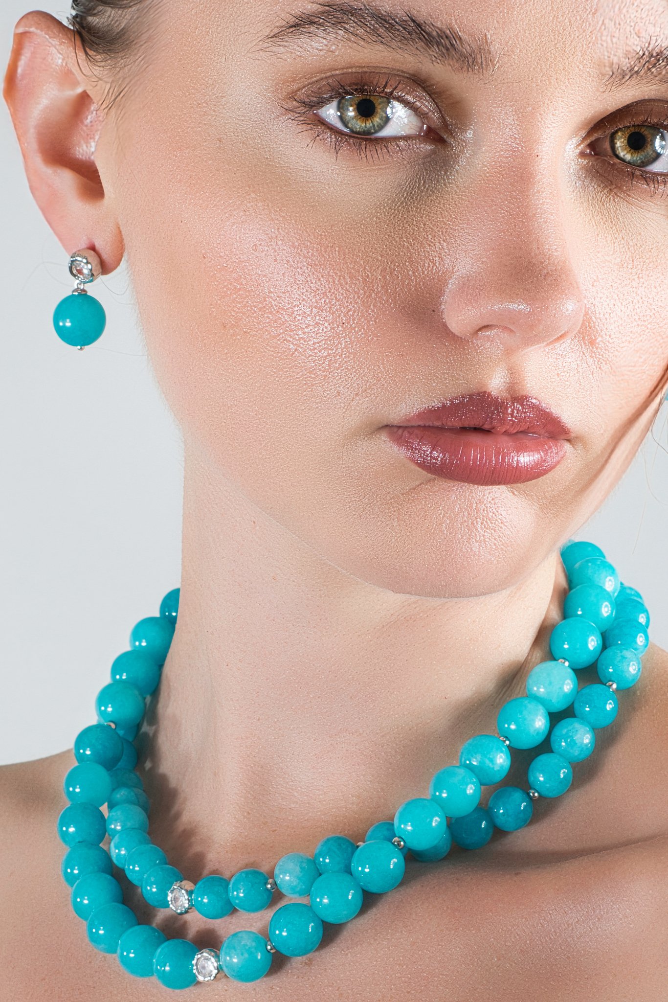 Close-up of a woman wearing turquoise jewelry, including earrings and a matching necklace, with a neutral background.