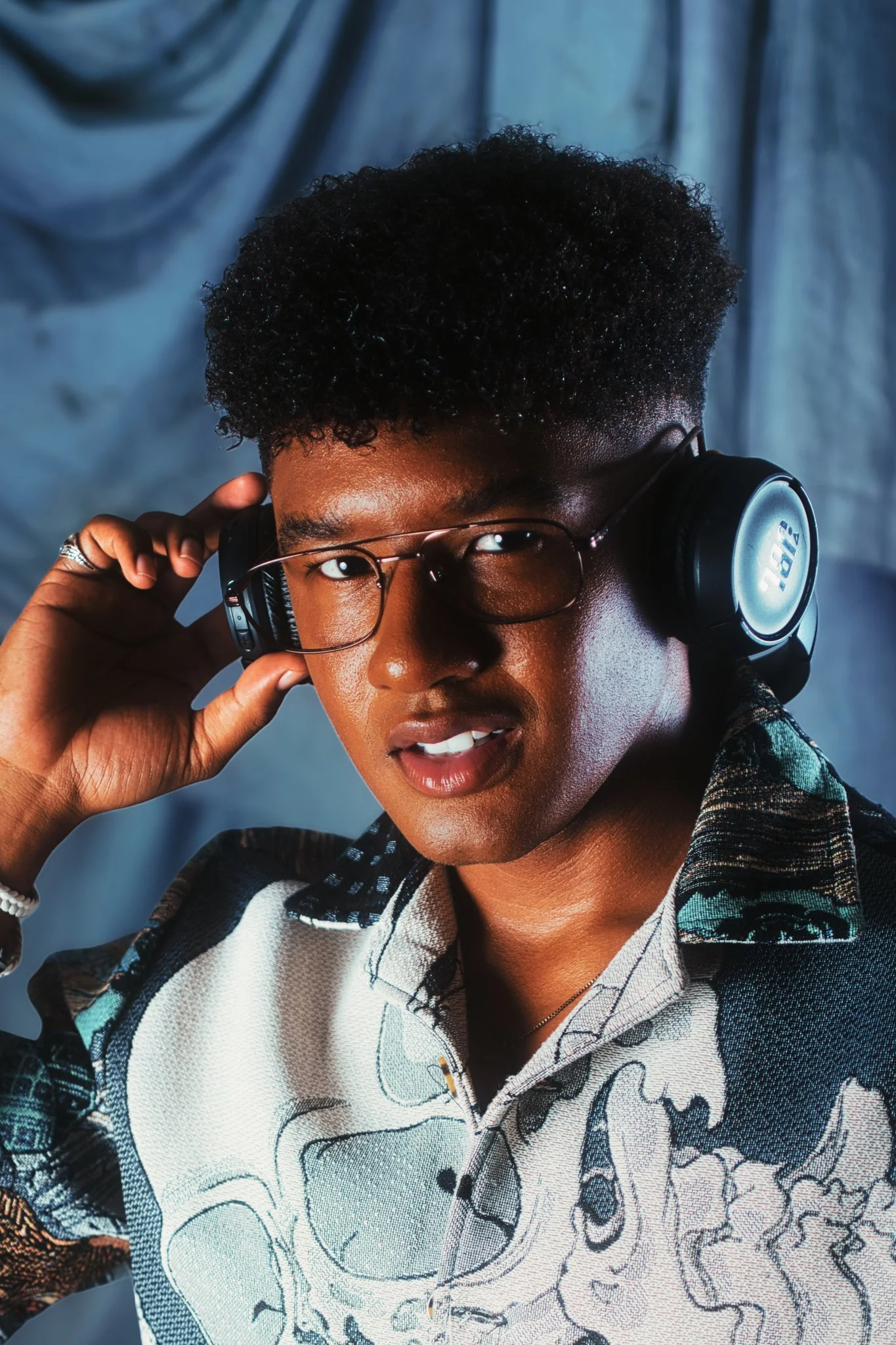 Young person with short curly black hair, glasses, and medium skin tone wearing headphones and a patterned shirt, looking at the camera.