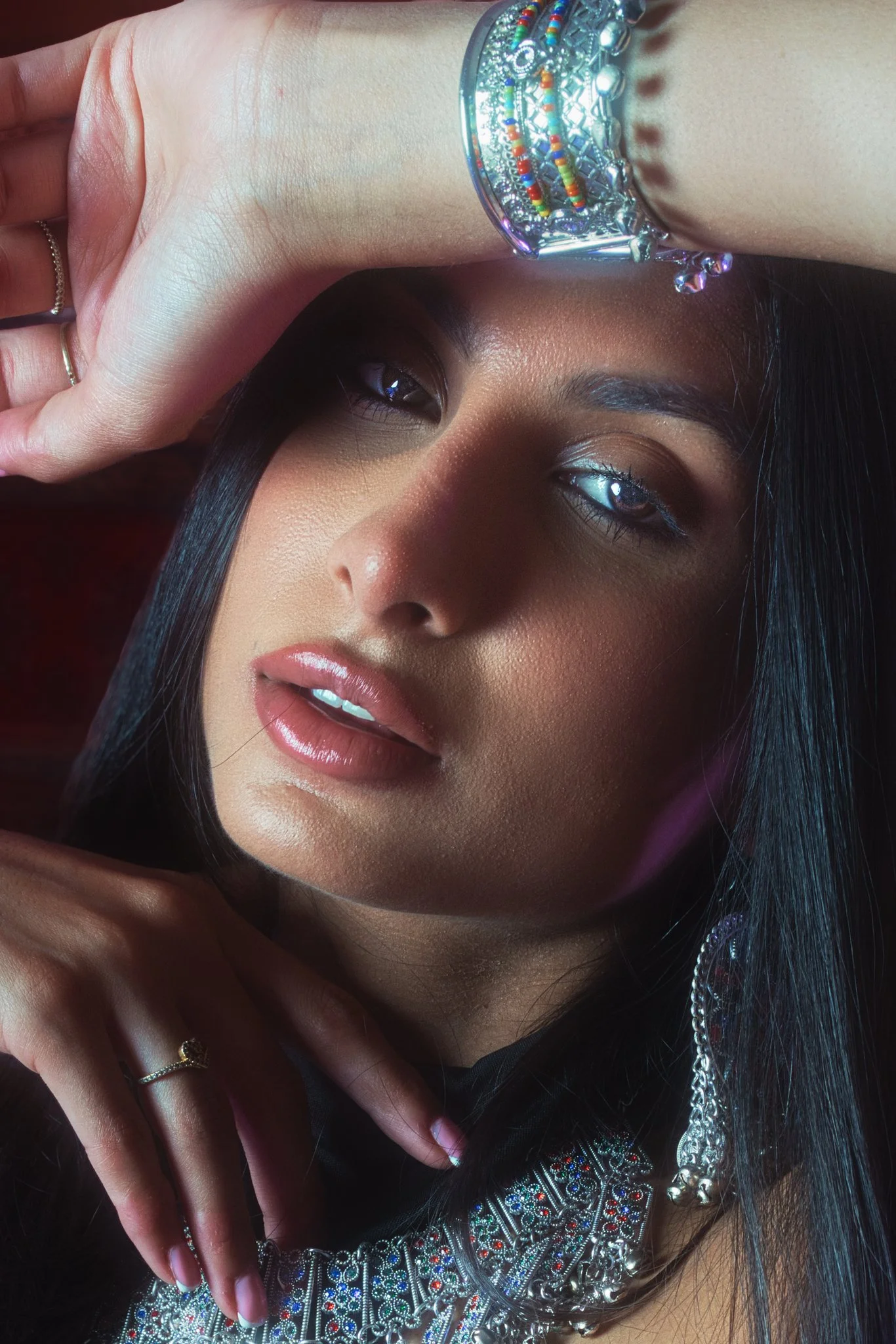 A woman with dark hair and brown skin is wearing colorful jewelry, including a large beaded bracelet and a necklace with intricate designs. She has her eyes half-closed and lips slightly parted, with her hand resting on her forehead, showcasing rings on her fingers.