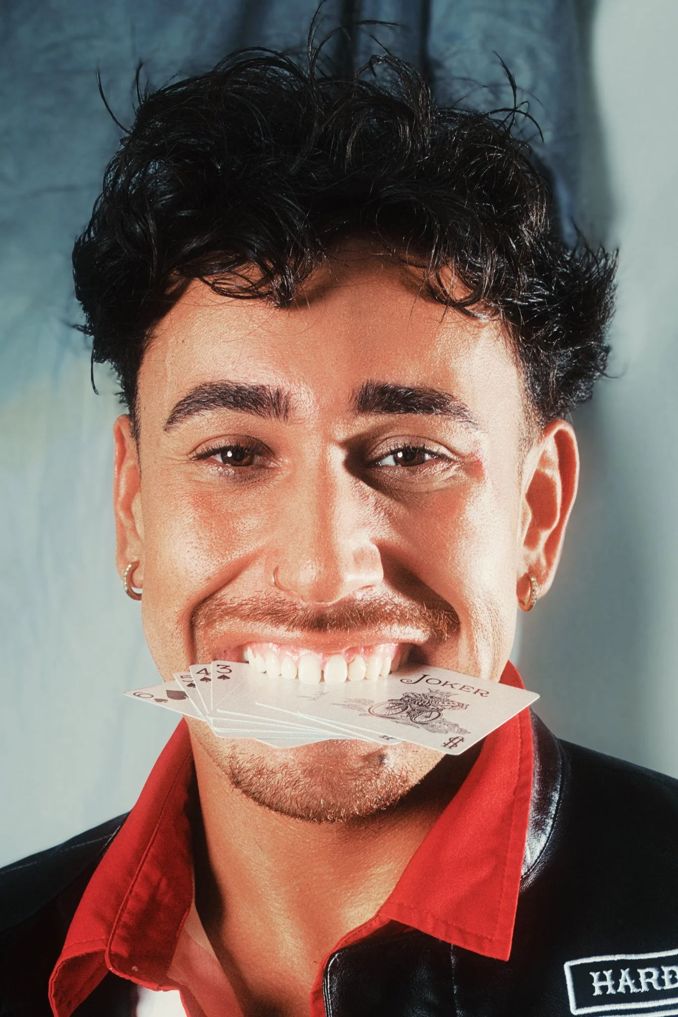 A smiling man with curly dark hair, slightly squinted eyes, earrings, and a goatee, holding a fan of playing cards in his mouth. He is wearing a red collared shirt and a black jacket with some white lettering on the chest.
