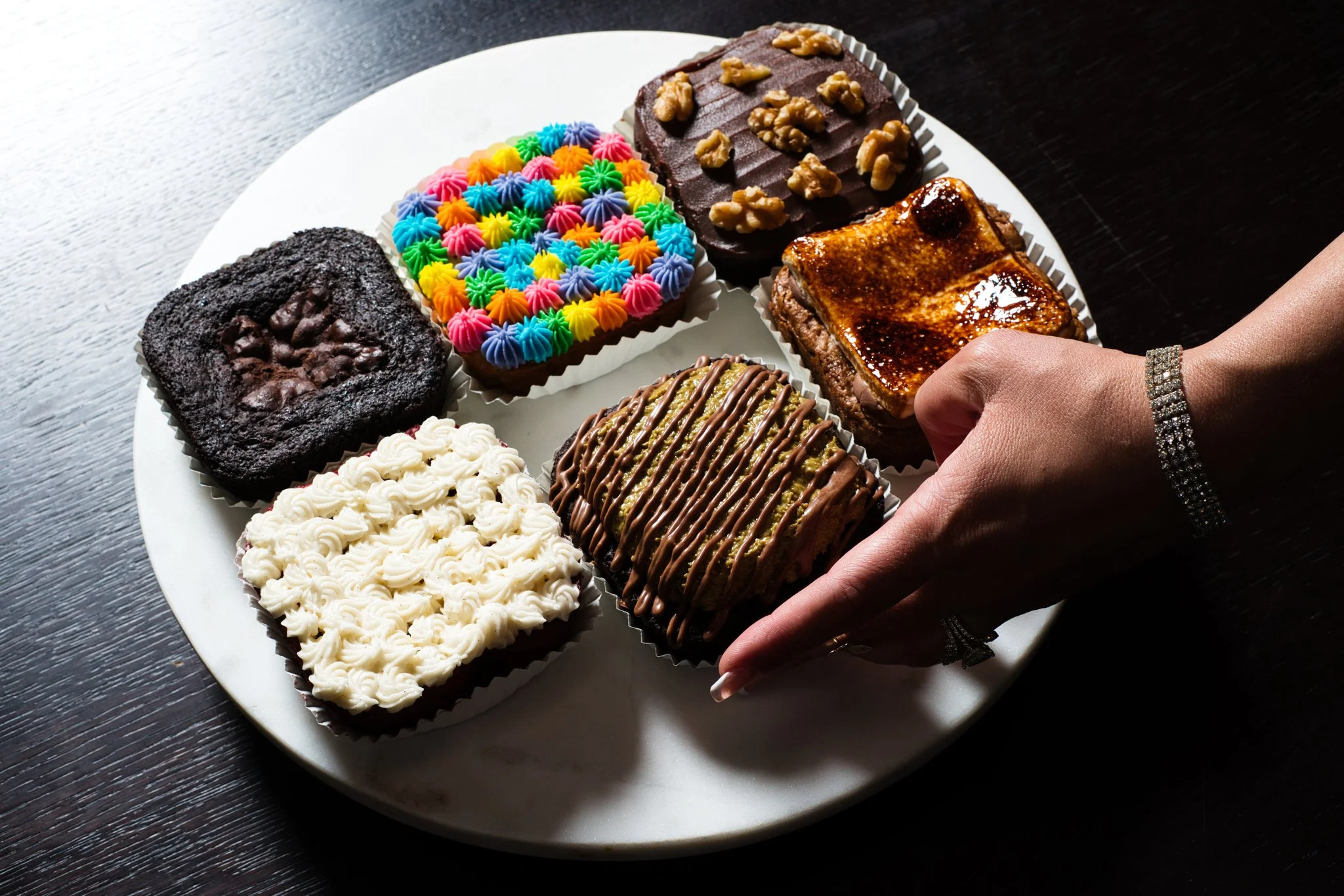 A white plate with five different types of colorful and decorated baked goods on a dark wooden surface, with a person's hand and wrist adorned with a diamond bracelet visible in the bottom right corner.
