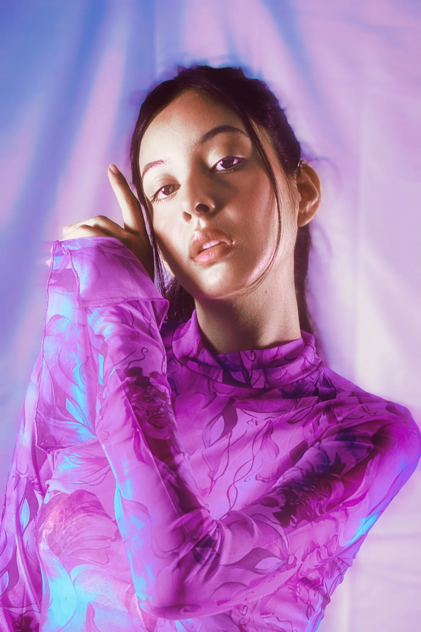 A young woman with dark hair in a ponytail, wearing a purple patterned long-sleeve top, looking at the camera with a neutral expression, against a soft purple and pink background.