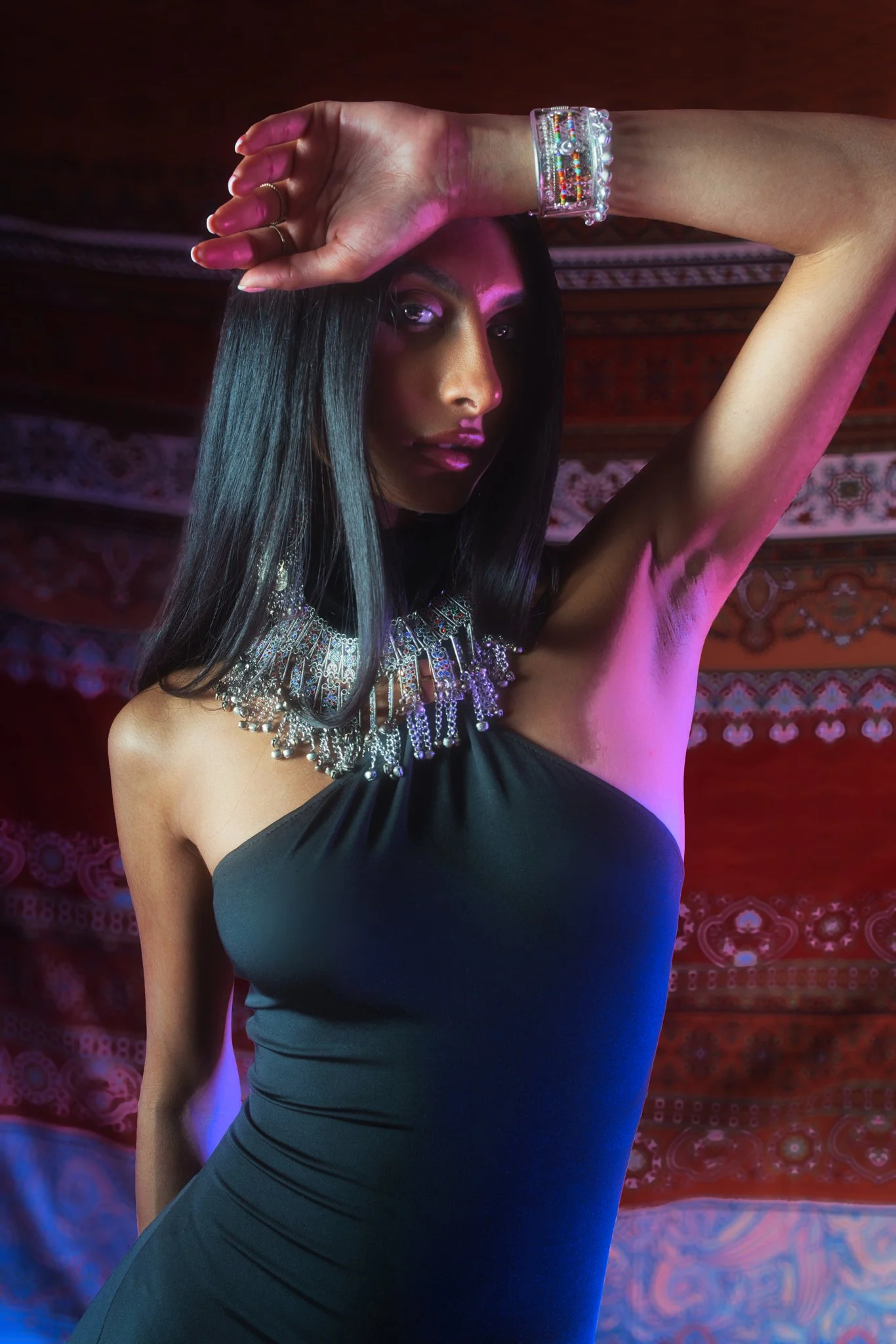 A woman with long black hair, wearing black clothing, silver jewelry, and a clear bracelet, posing with her arm across her forehead in front of a vibrant red and purple patterned background with colorful lighting