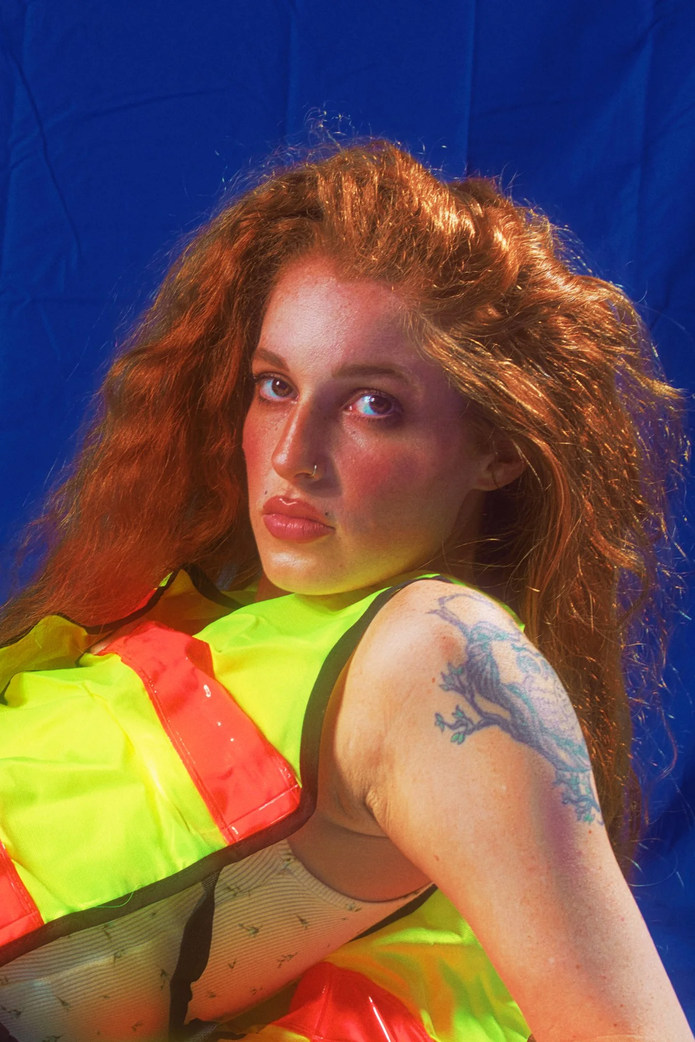 A young woman with red hair, blue eyes, and tattoos on her arm, wearing a neon yellow vest and striped pants, taking a selfie against a blue background.