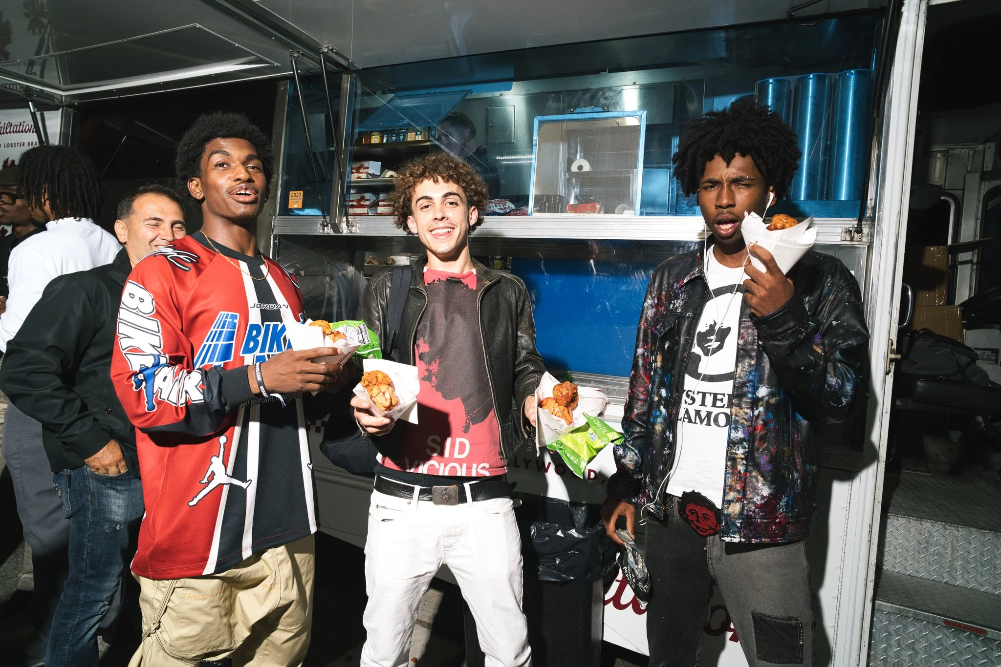 Group of five young men standing in front of a food truck, holding pieces of fried chicken in paper trays, smiling and enjoying their meal.
