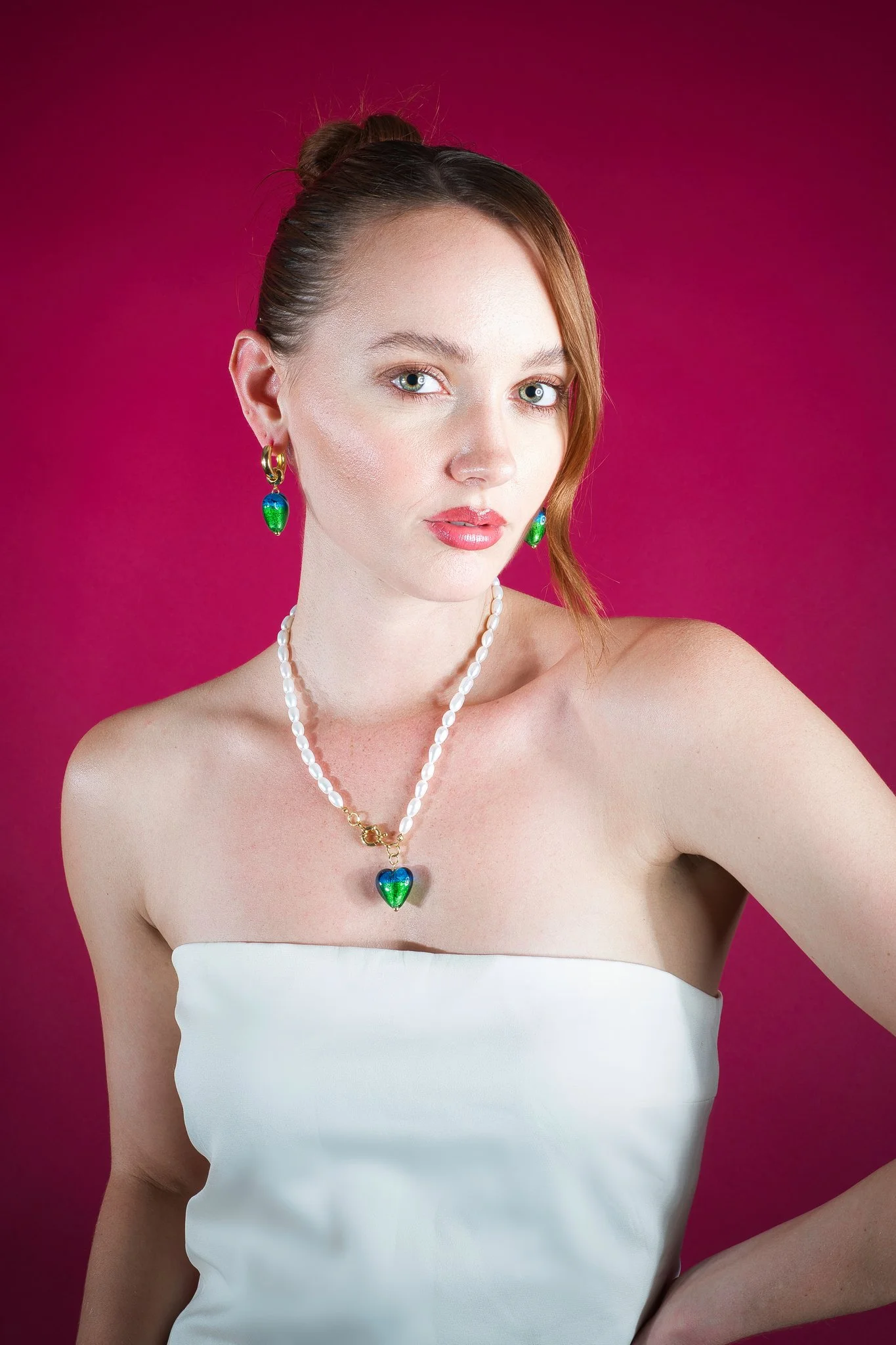A woman with fair skin, light makeup, blue eyes, and long brown hair styled with one side tucked back and the other side hanging over her shoulder, wearing pearl jewelry with green and blue heart-shaped pendants, against a pink background.