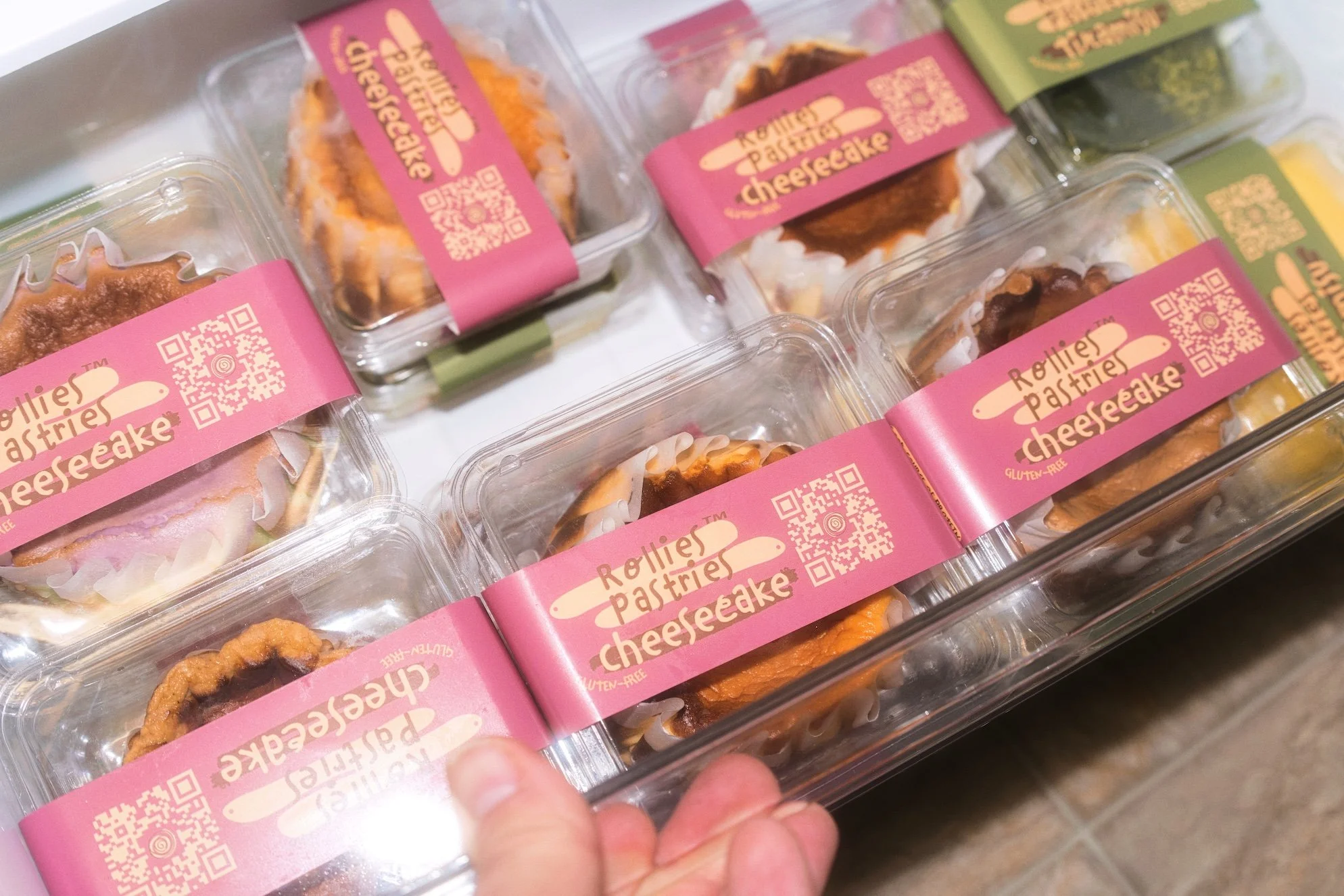 Multiple clear plastic containers filled with gluten-free cheesecakes labeled 'Roller Pastries Cheesecake' with pink labels and QR codes, stacked closely together.