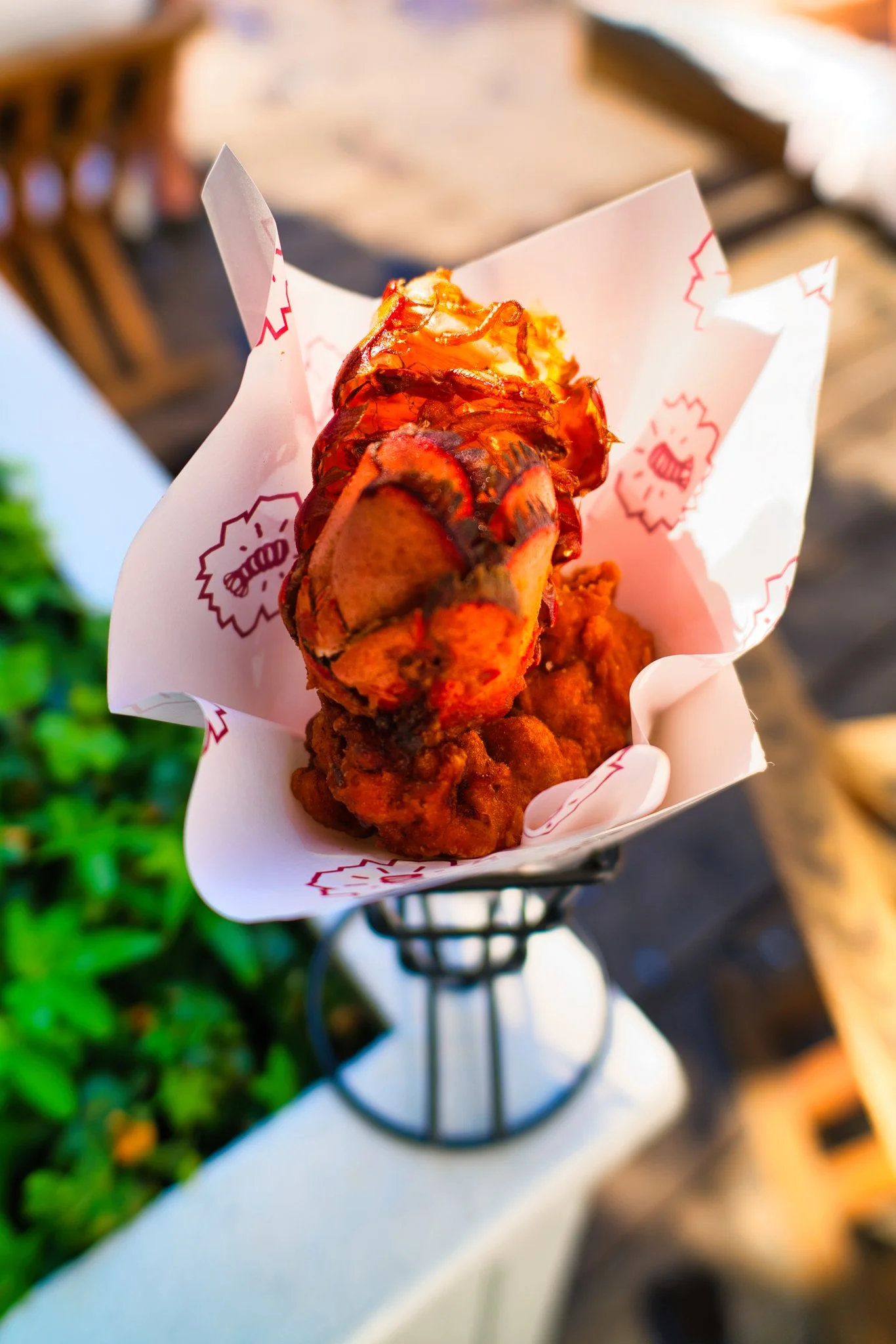 A person holding a deep-fried seafood snack, possibly a lobster roll or seafood battered item, wrapped in white paper with red graphics, in an outdoor setting.