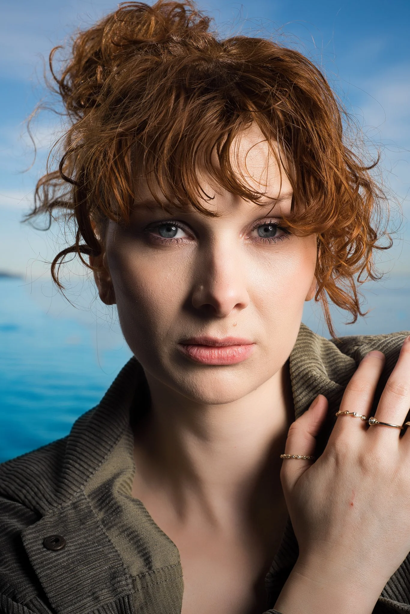 Close-up portrait of a woman with curly red hair and blue eyes, wearing a green jacket, standing outdoors near water with a blue sky in the background.