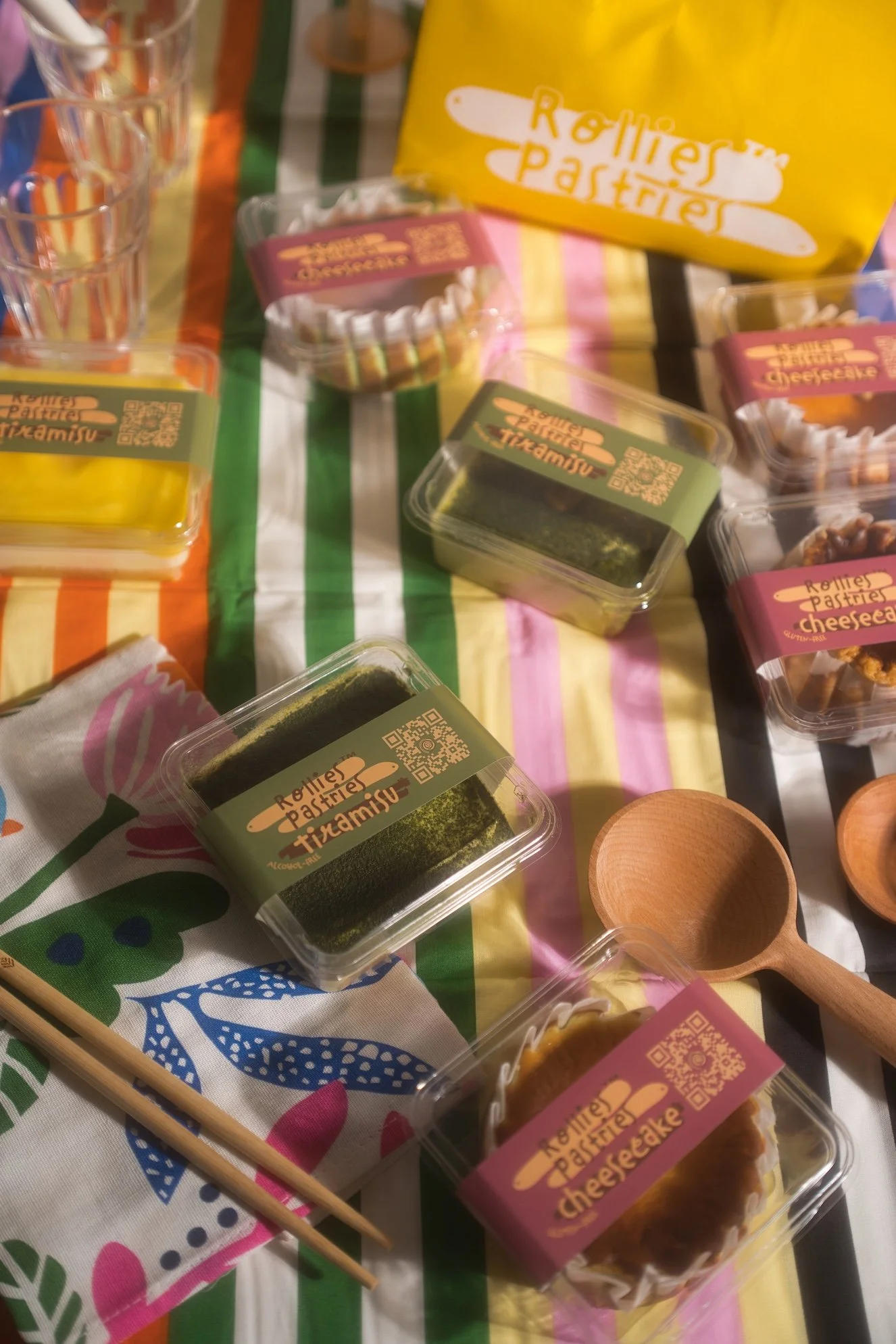 Assorted small desserts including cheesecakes and tiramisu in clear plastic containers, placed on a colorful tablecloth with a wooden spoon and chopsticks nearby.