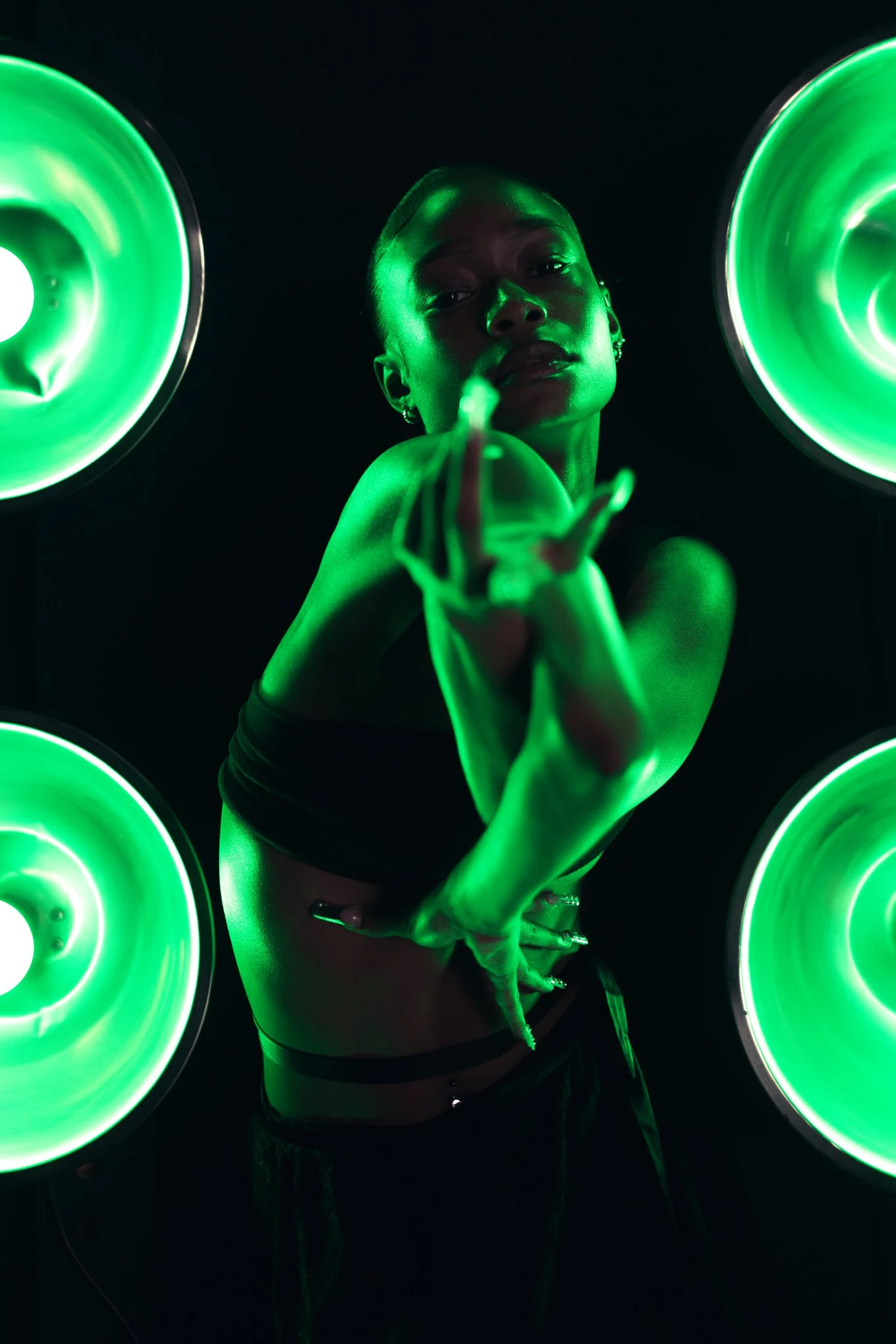 A woman with dark skin posing in front of four large green lights, with her hand extended toward the camera, and a confident expression.