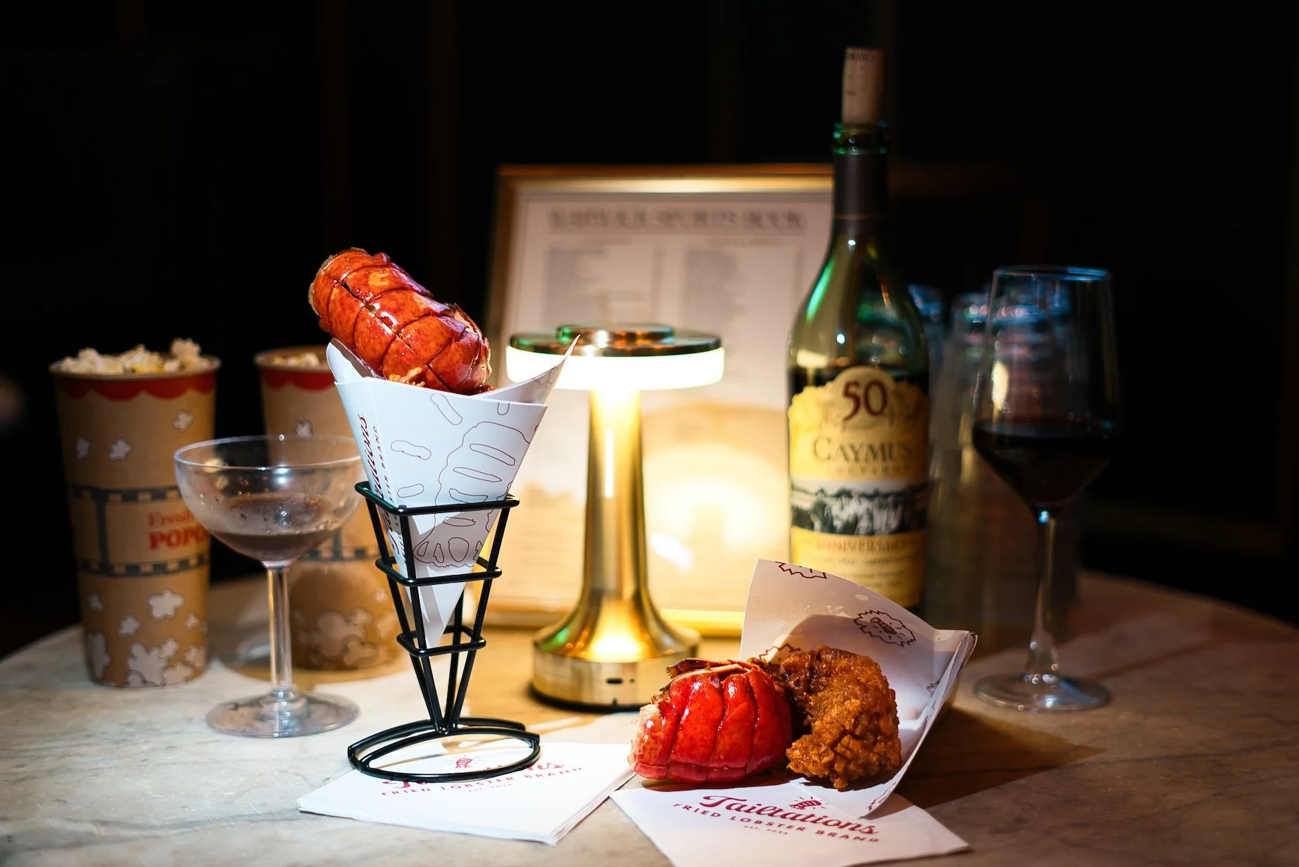 A close-up of a lobster roll in a white paper cone, fried chicken pieces on a white paper, a glass of red wine, a bottle of Calvet wine, and popcorn containers in the background, set on a wooden table with a warm lit ambient.