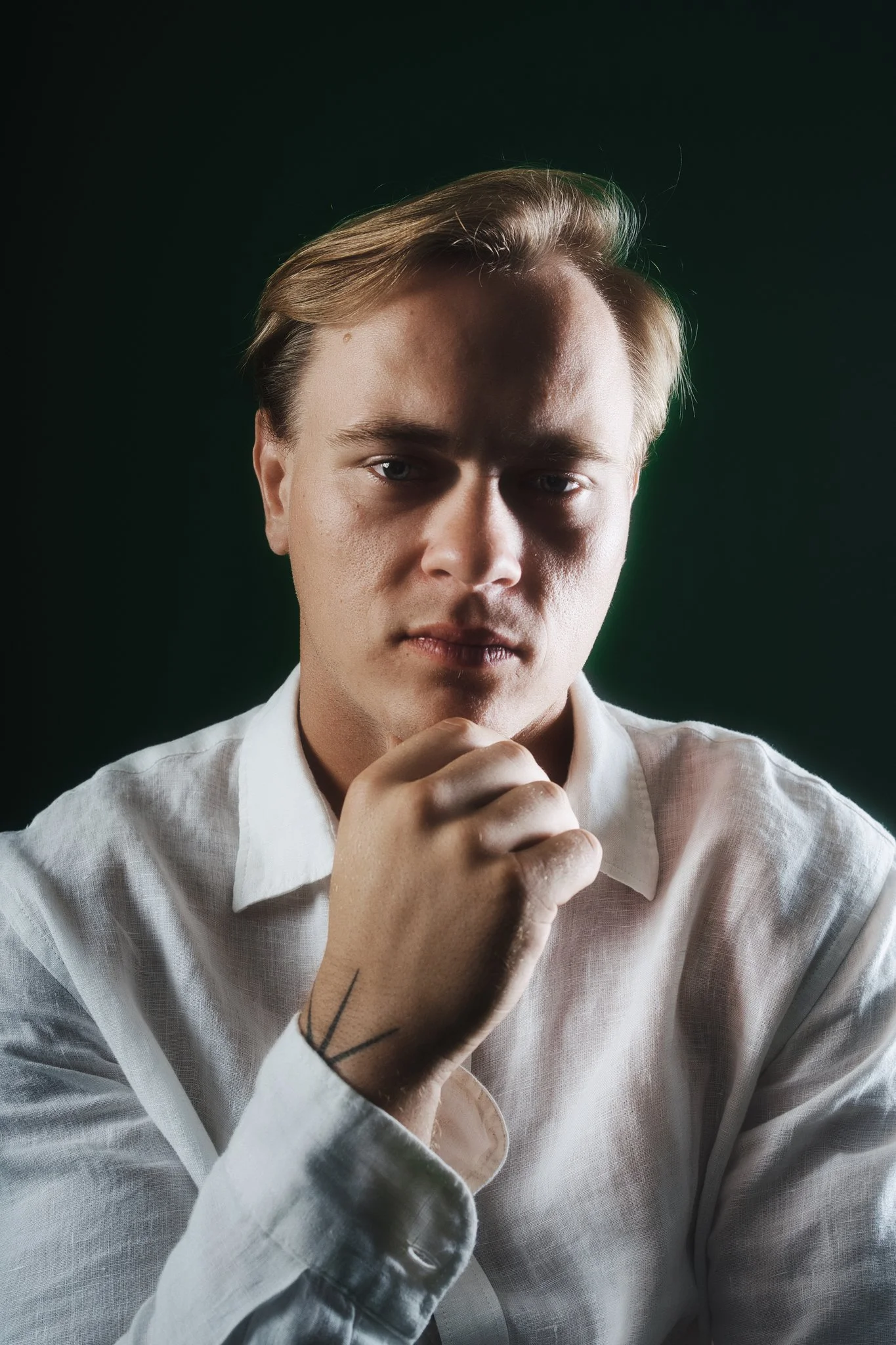 A young man with blonde hair styled to the side, wearing a white shirt, looking directly at the camera with a serious expression, holding his chin with his hand, against a dark background.